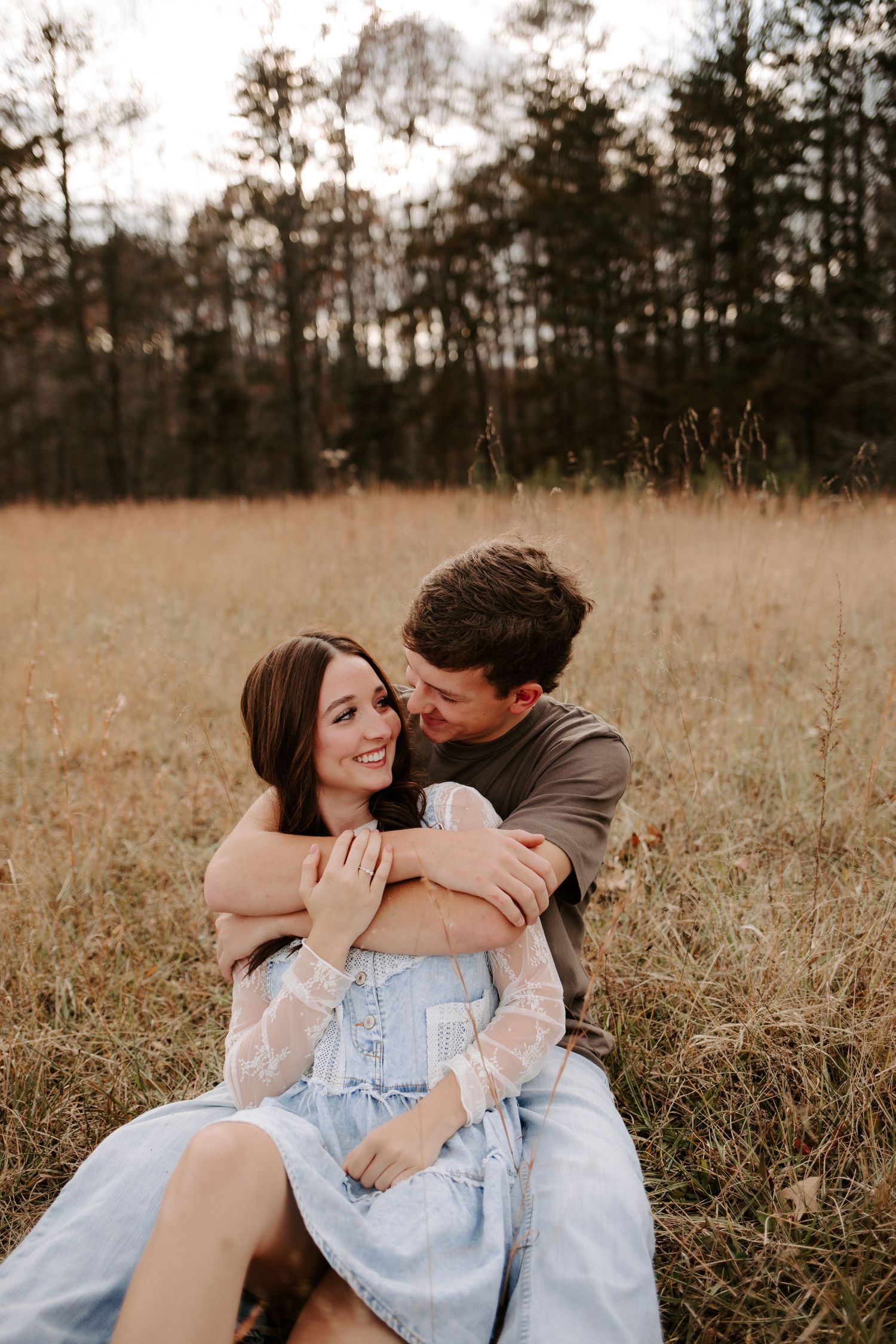 A couple sits together in a golden field, the man embracing the woman in a blue lace dress as she smiles warmly.