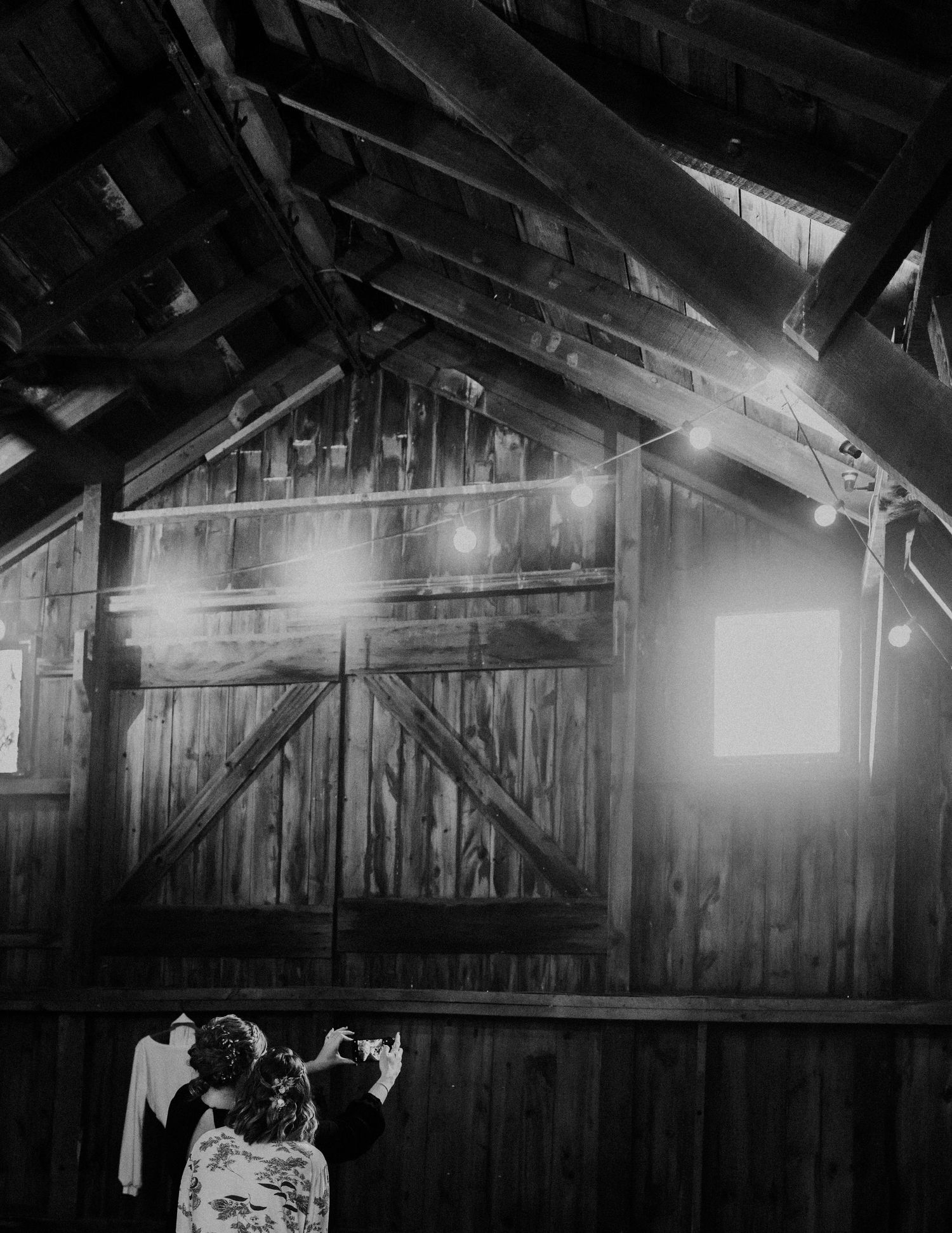 Black and white photo of rustic barn interior with light streaming through wooden beams.