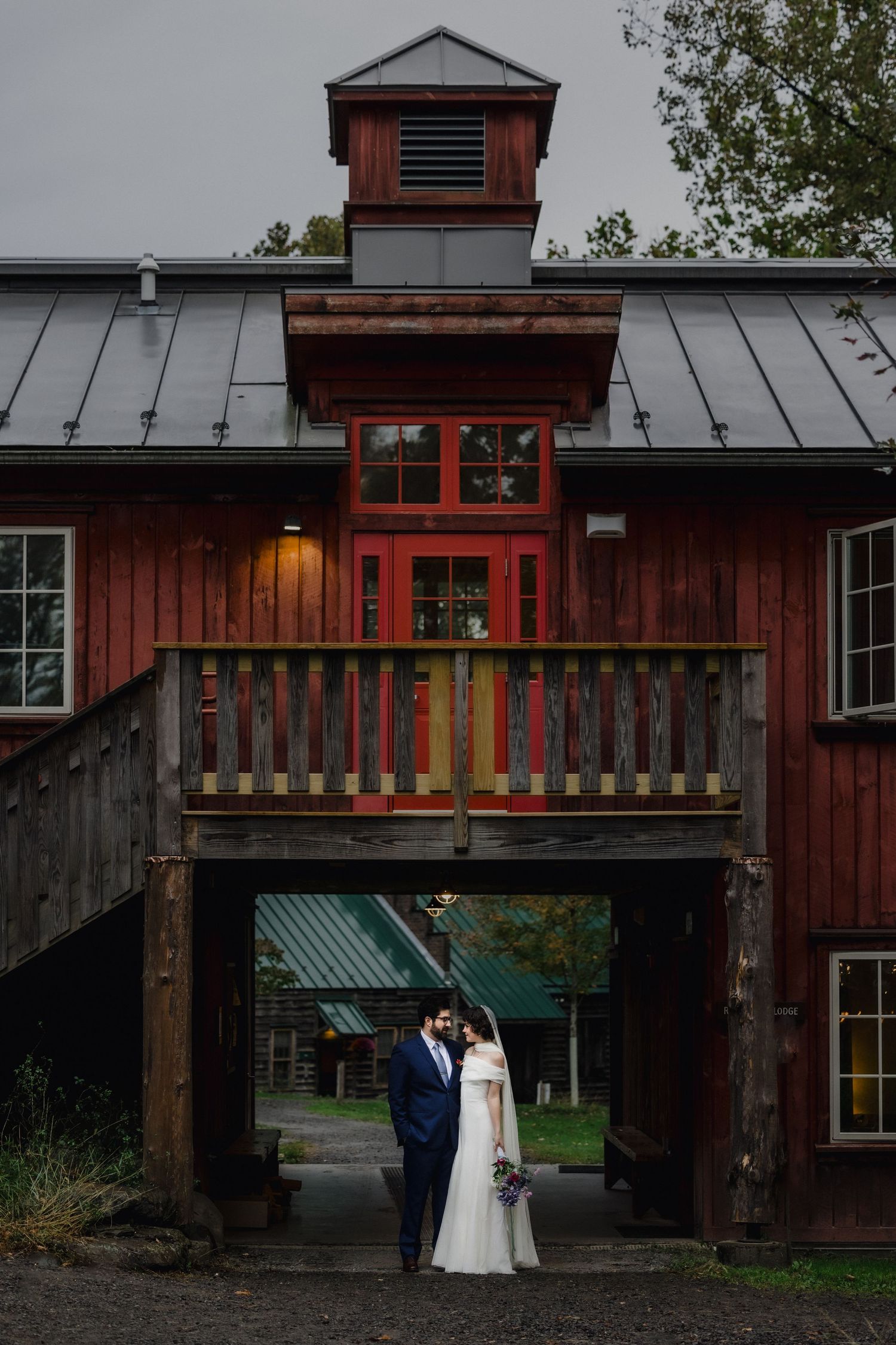 A wedding couple poses under a rustic red barn entrance with wooden railings and metal roof.