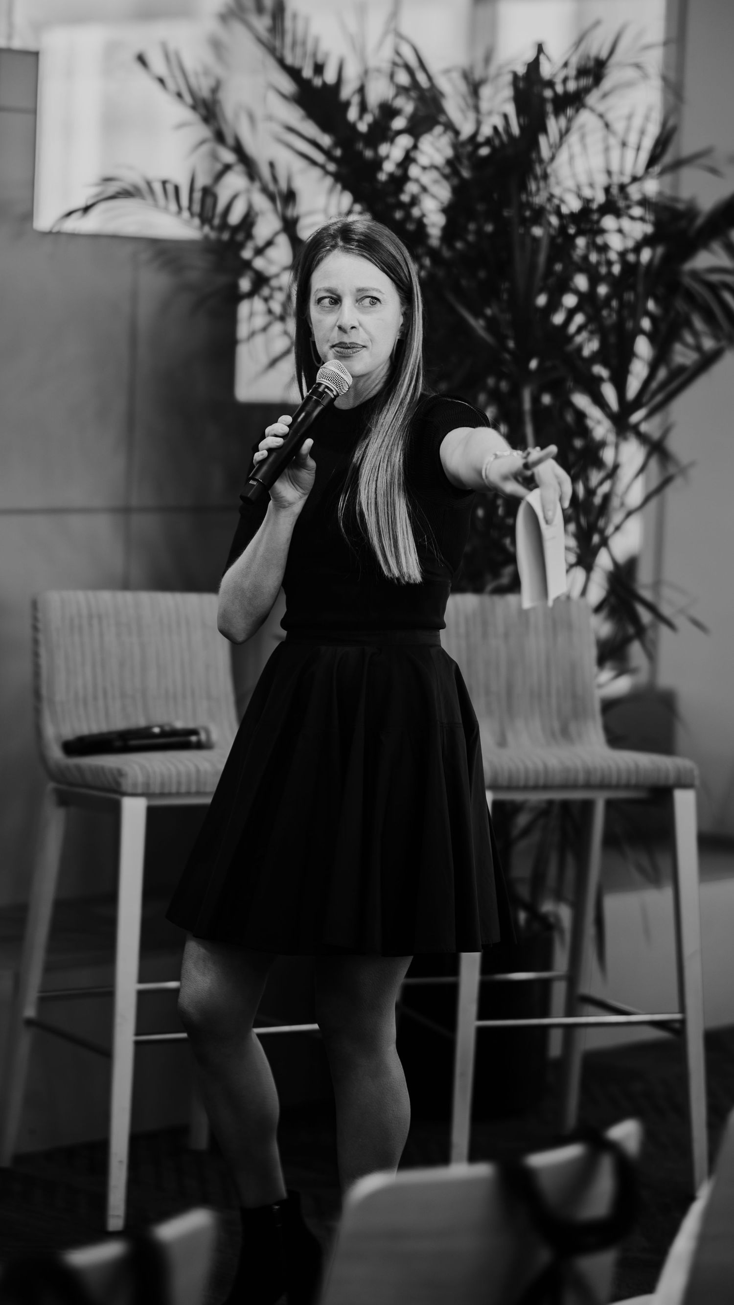 Presenter in black dress speaking expressively against palm plant backdrop.