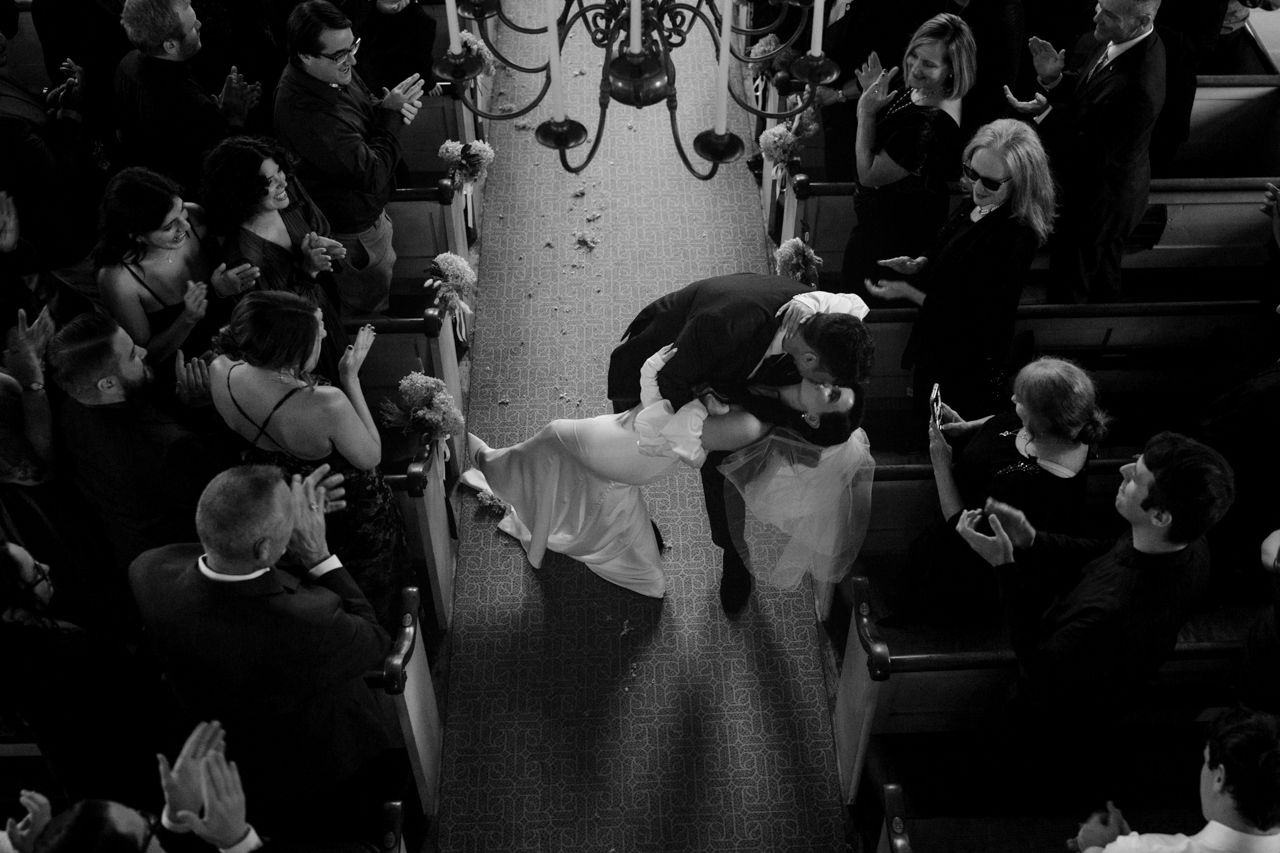 Black and white photo of wedding guests dancing under a chandelier in a crowded reception hall.