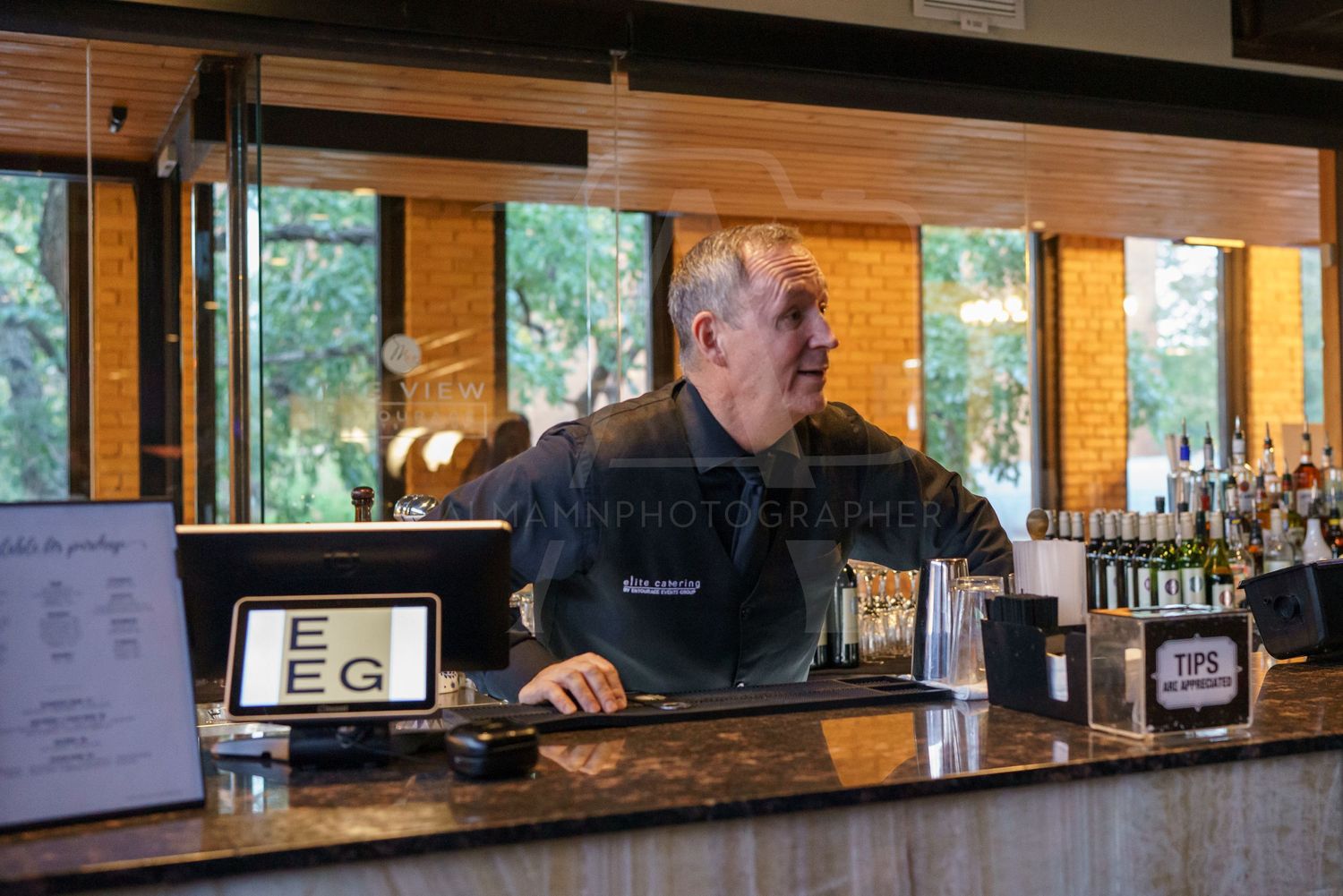 Bar interior with wooden decor and large windows showing bartender at work station.
