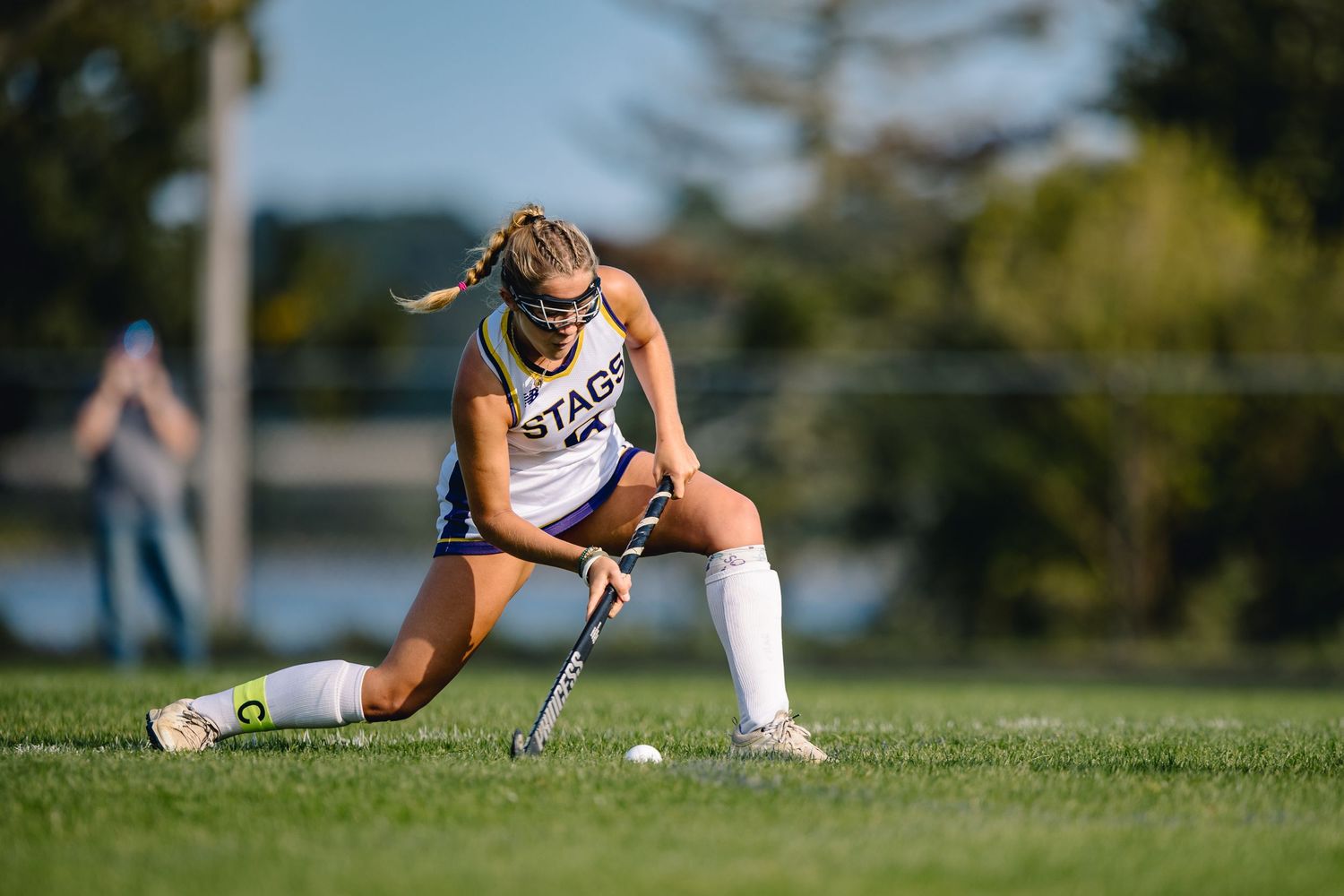 Field hockey player in white uniform kneeling on grass with stick and ball.