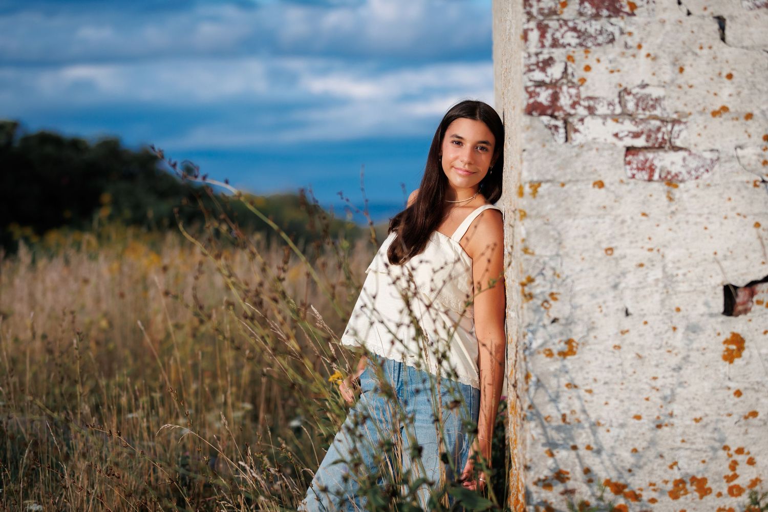 Senior HS Student in a sleeveless floral dress leans against a rustic white brick wall with tall grass and blue sky in the background.