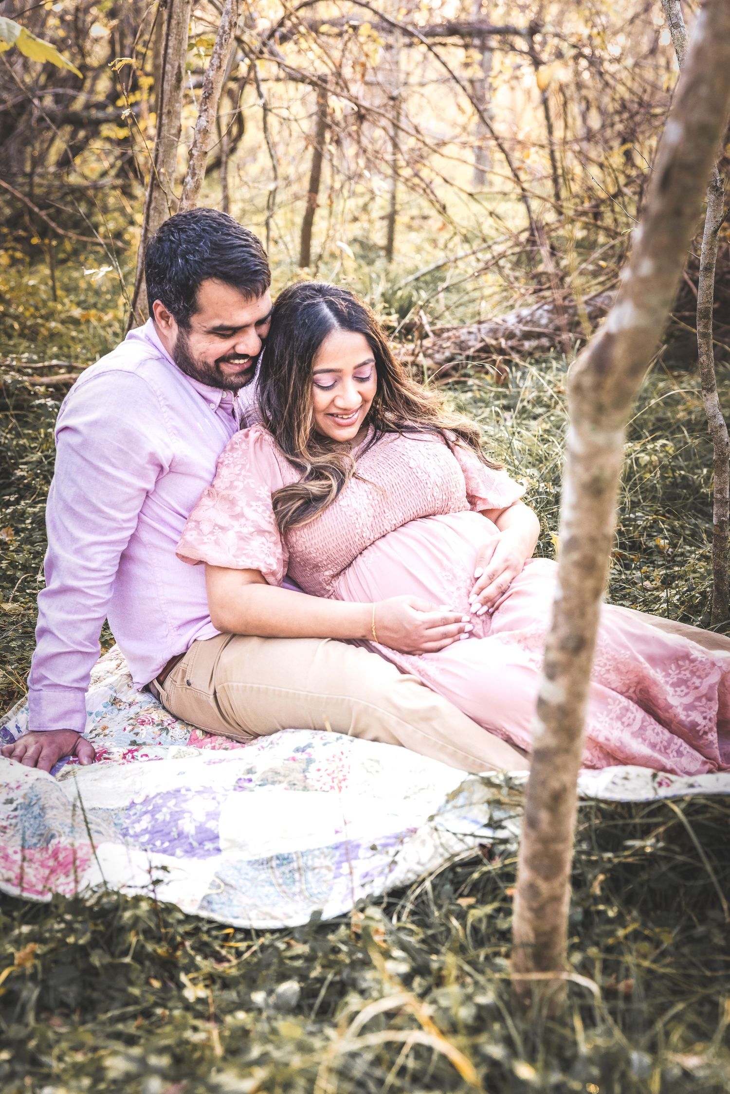 A couple shares tender moments while relaxing on a blanket in a sunlit wooded area during an outdoor photo session.
