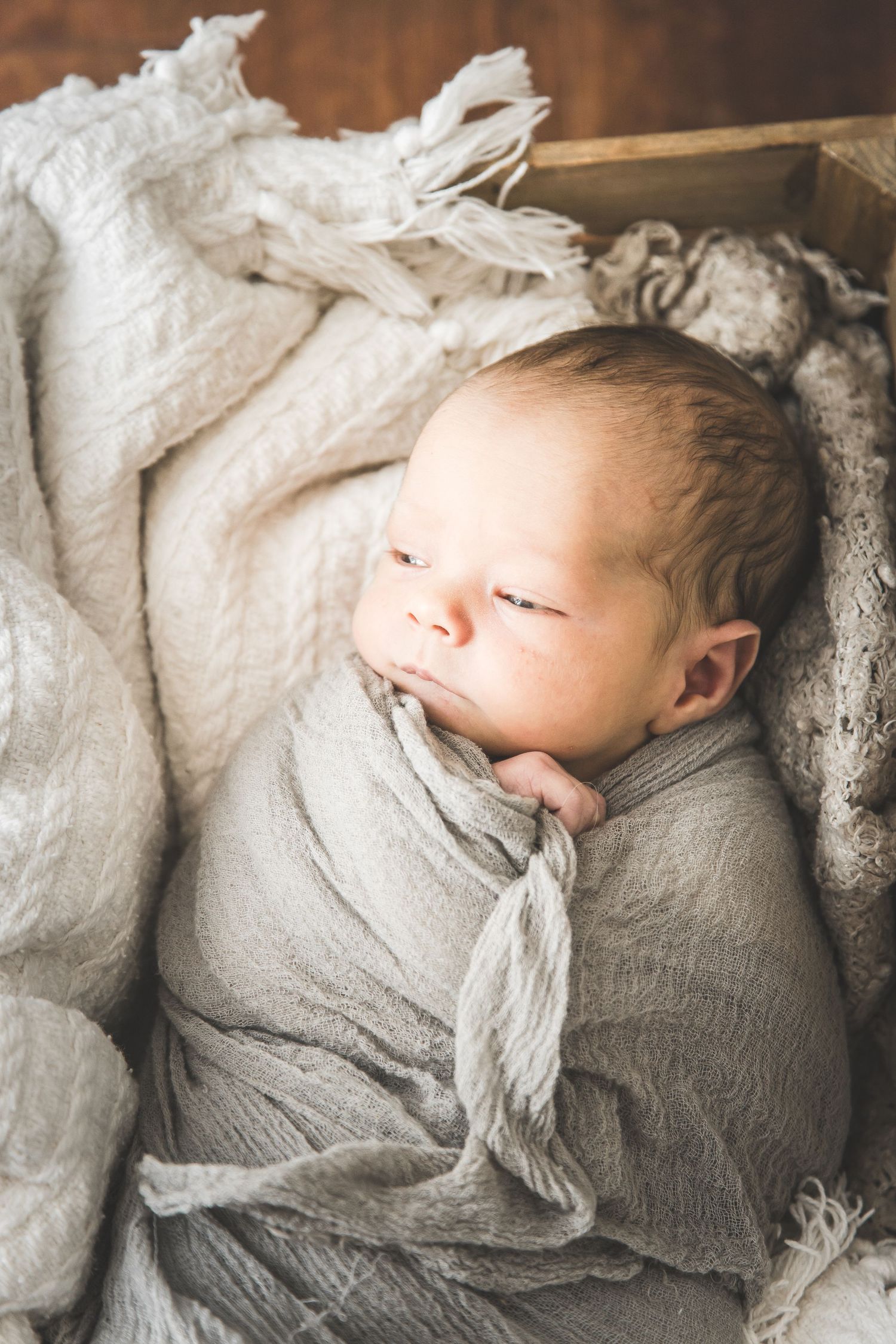 A sleeping newborn wrapped in soft gray and white linens rests peacefully in a wooden box.
