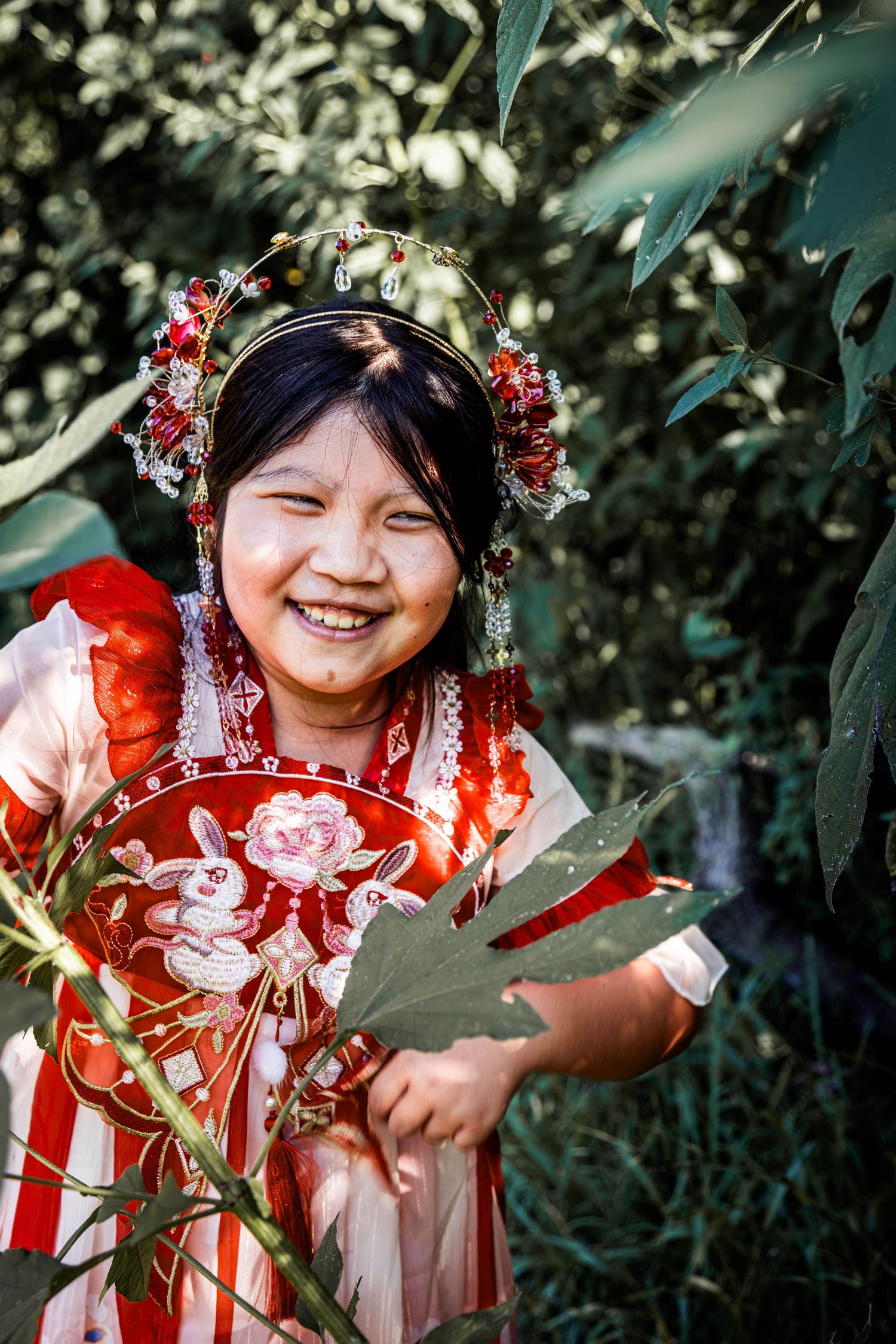 Smiling child in traditional Asian attire with floral headdress standing among green foliage.  Pearland Family Photographer