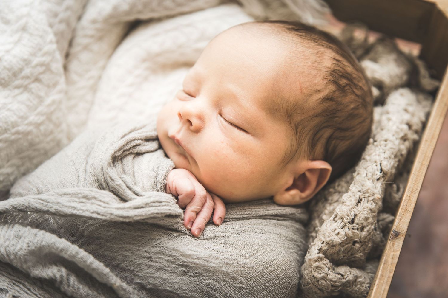 A peaceful sleeping newborn baby wrapped in soft gray swaddle blanket rests on textured bedding.  Pearland Family Photographer