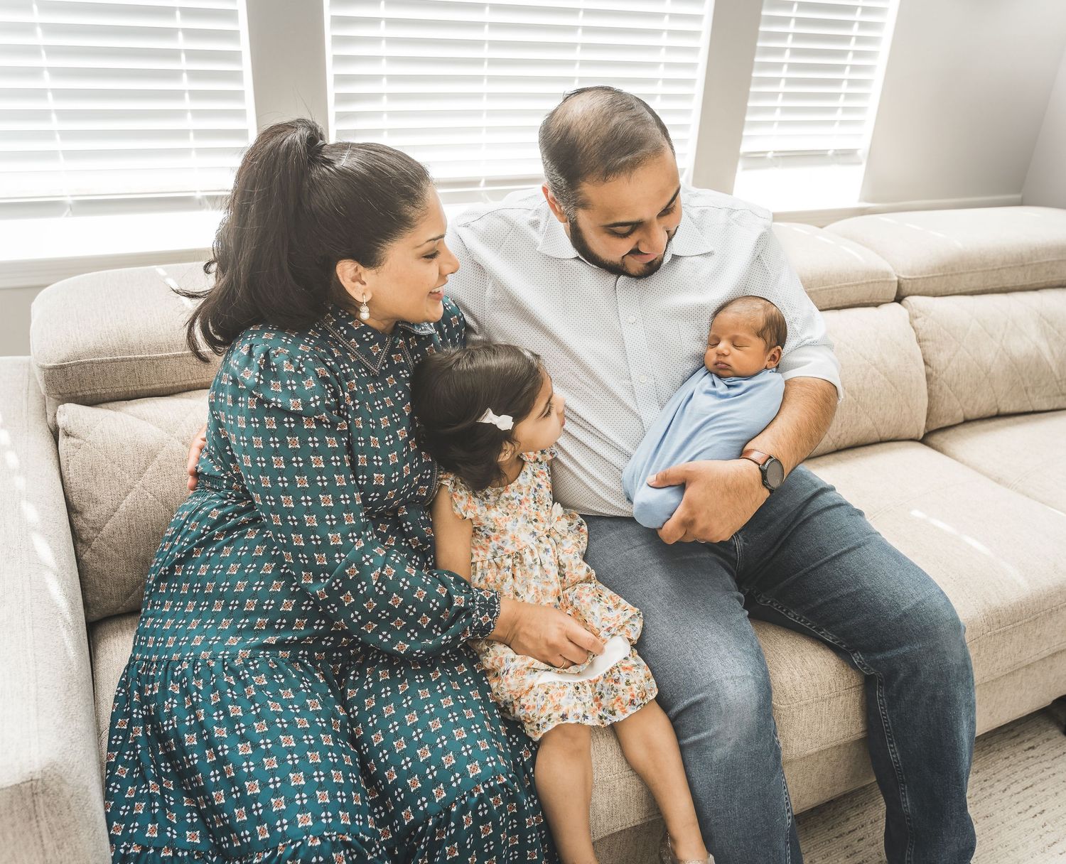 A family sits together on a gray couch in a bright living room with window blinds in the background.