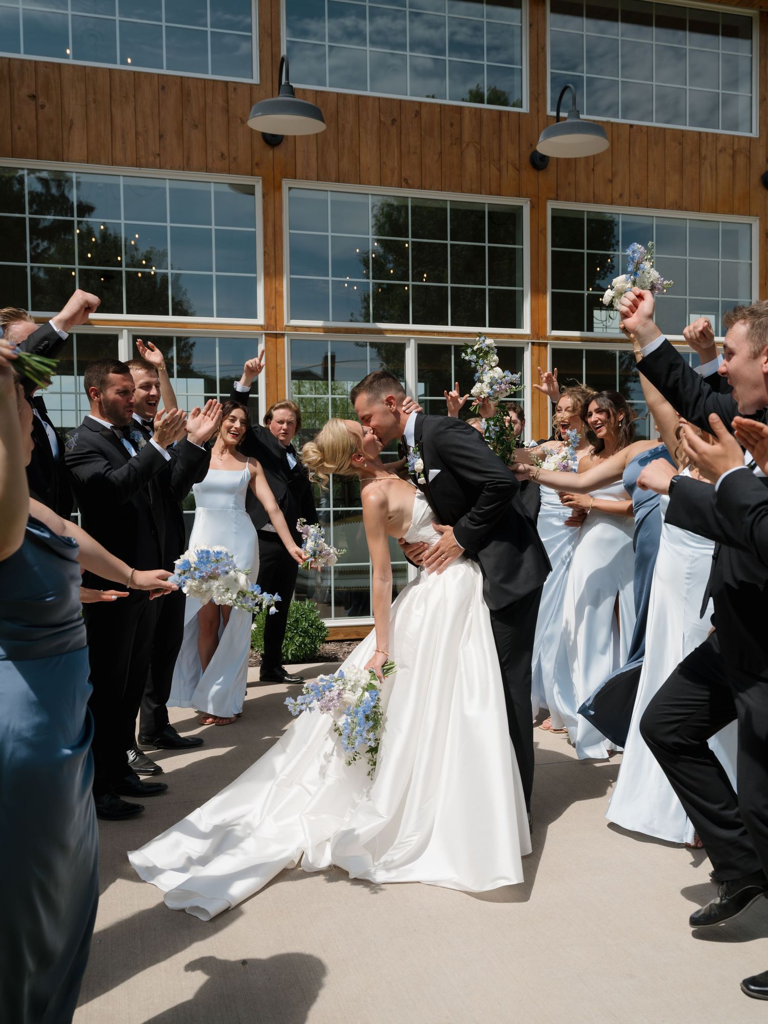 Bride and groom share a kiss surrounded by cheering wedding party outside a modern venue on a sunny day.
