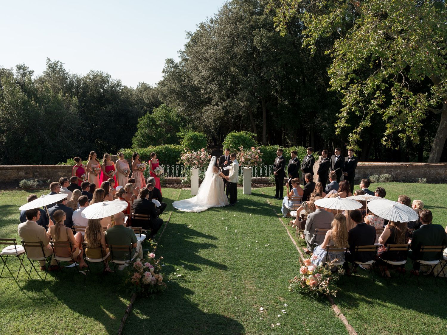 Outdoor wedding ceremony on lush green lawn with guests seated in rows, bride in white gown, surrounded by tall trees.