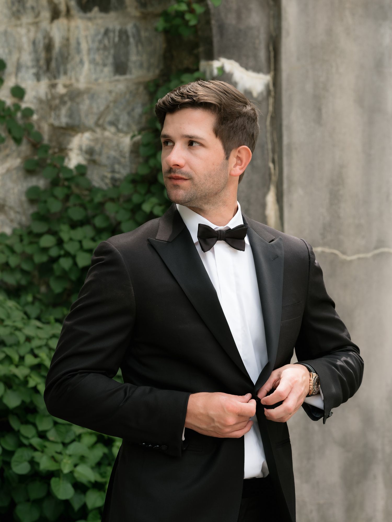 Groom in black tuxedo with bow tie buttoning jacket outdoors.