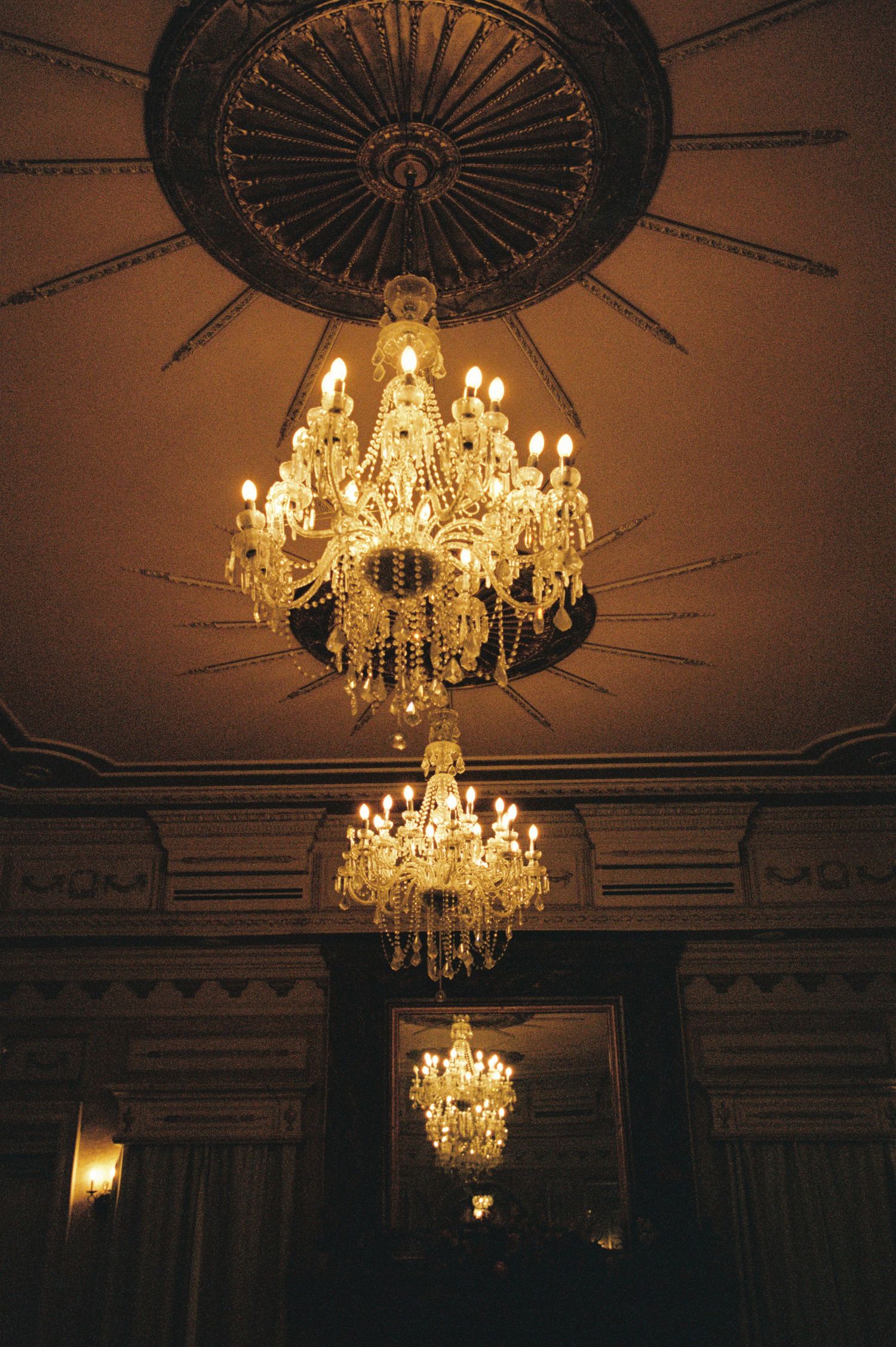 Elegant crystal chandeliers hanging from ornate ceiling medallion in a grand historic room with warm candlelight glow.