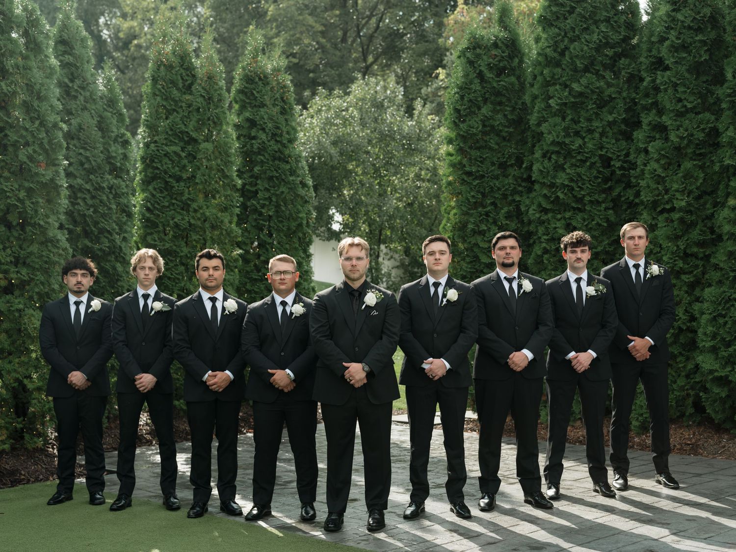 Wedding party members in black suits stand in line on stone steps surrounded by lush greenery.