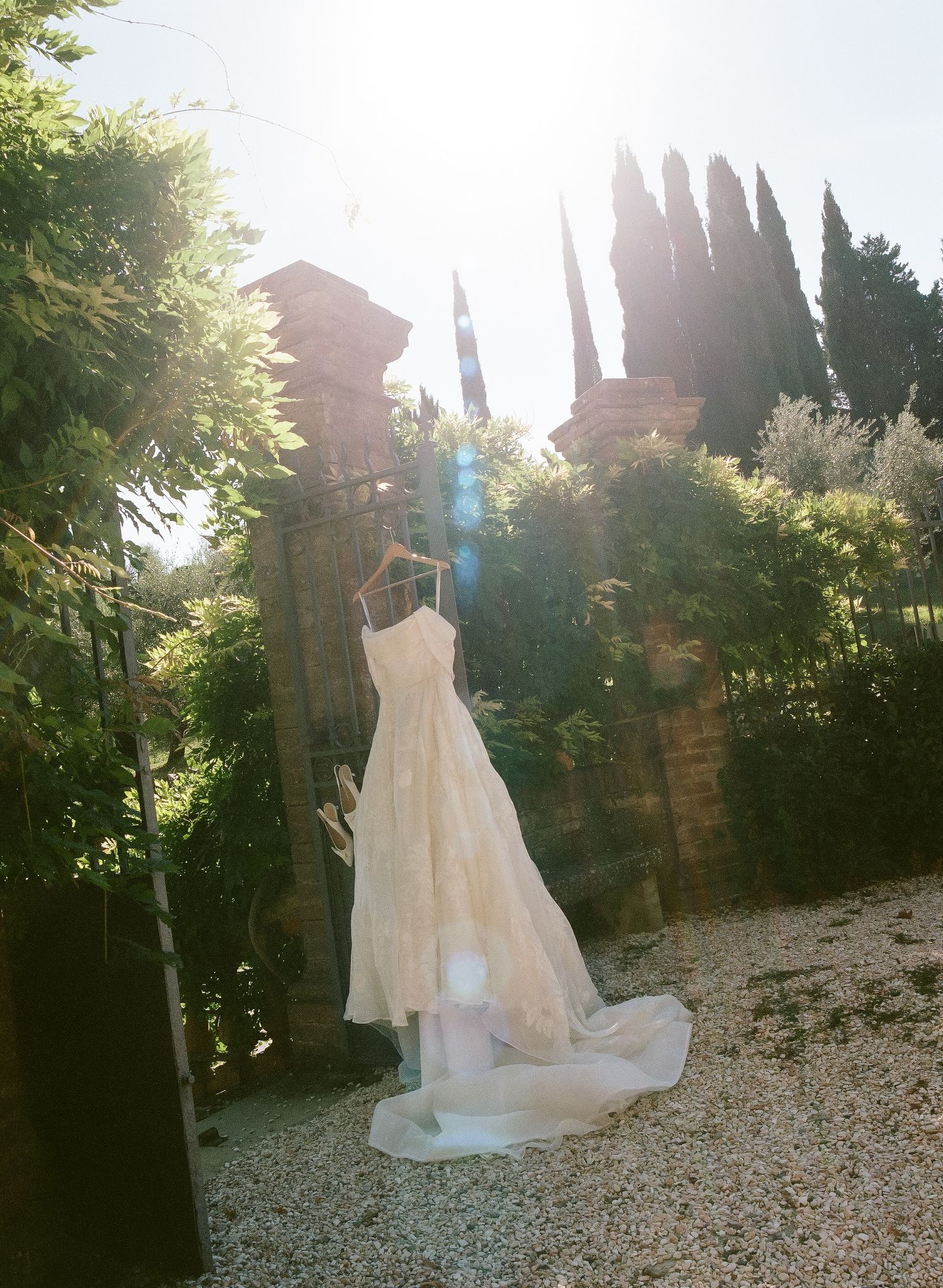 Elegant wedding dress hanging outdoors in a sunlit Italian garden with cypress trees and stone pillars.