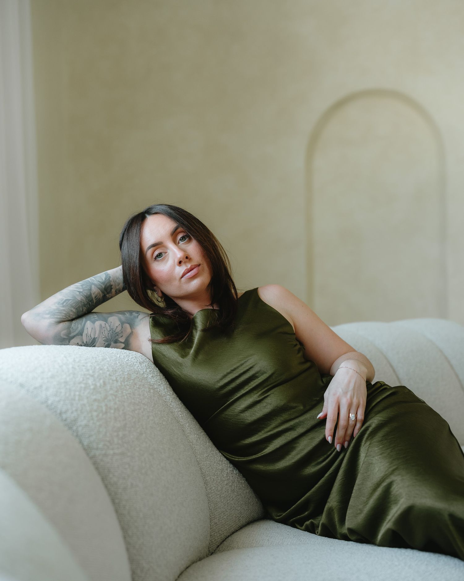 Woman in olive satin dress with tattoo sleeve reclines on white boucle sofa against neutral arched wall.