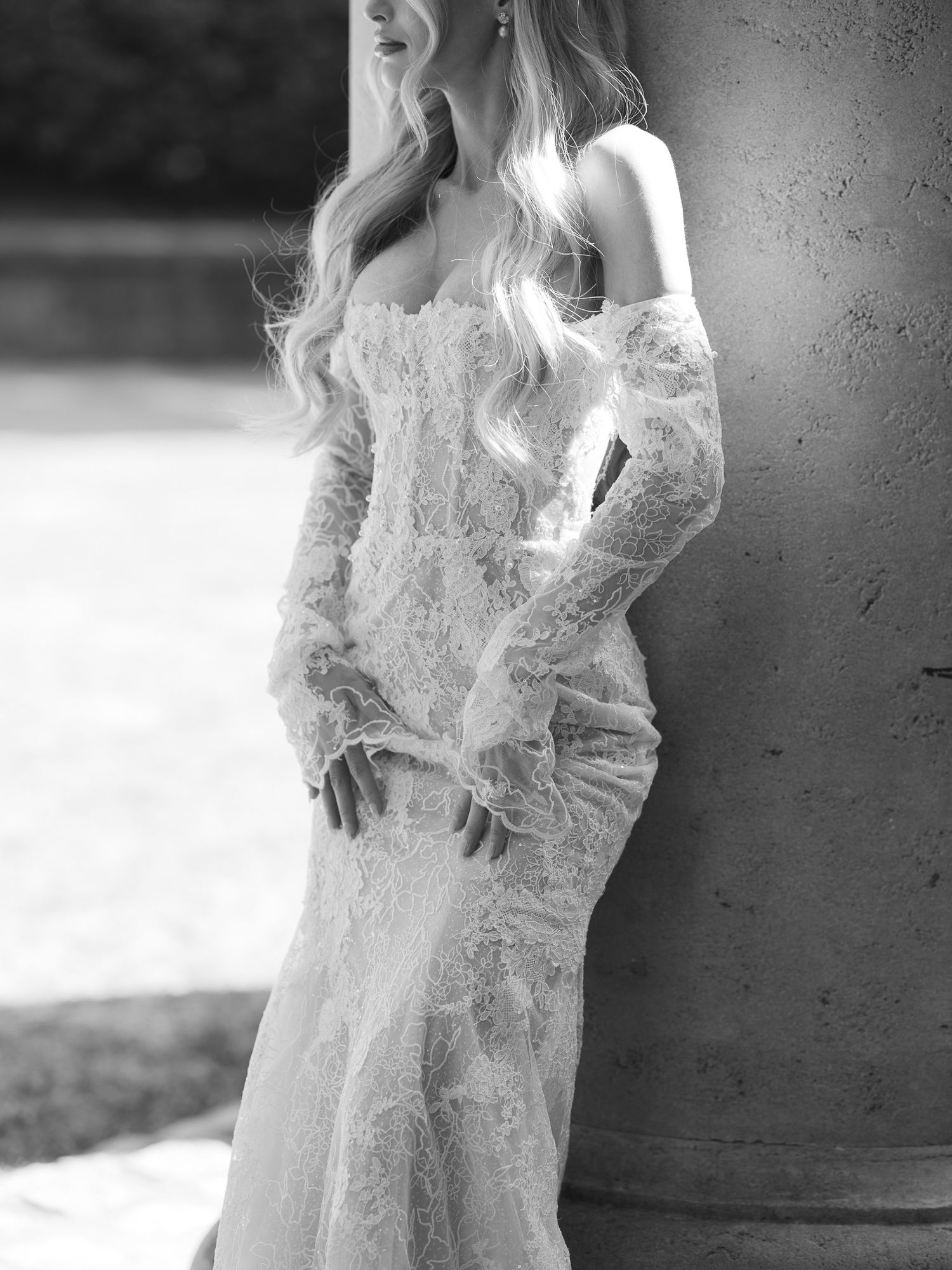 Black and white photo of a bride in a lace strapless gown with sheer gloves, leaning against a stone column.