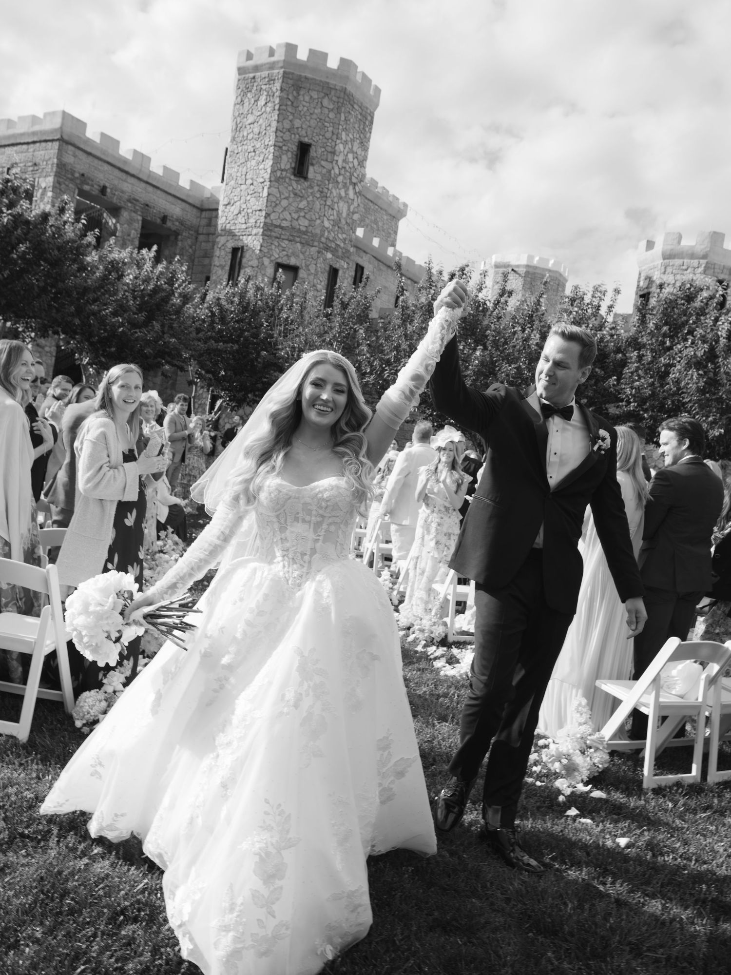 Black and white photo of newlyweds celebrating recessional at outdoor castle wedding ceremony with guests cheering.