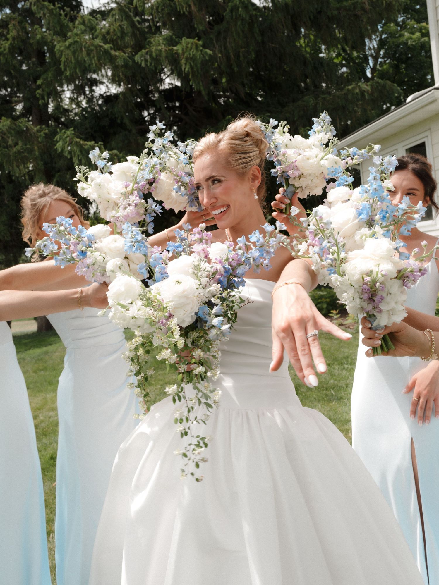 Bride in white gown with cascading blue and white floral bouquet surrounded by bridesmaids in light blue dresses outdoors.