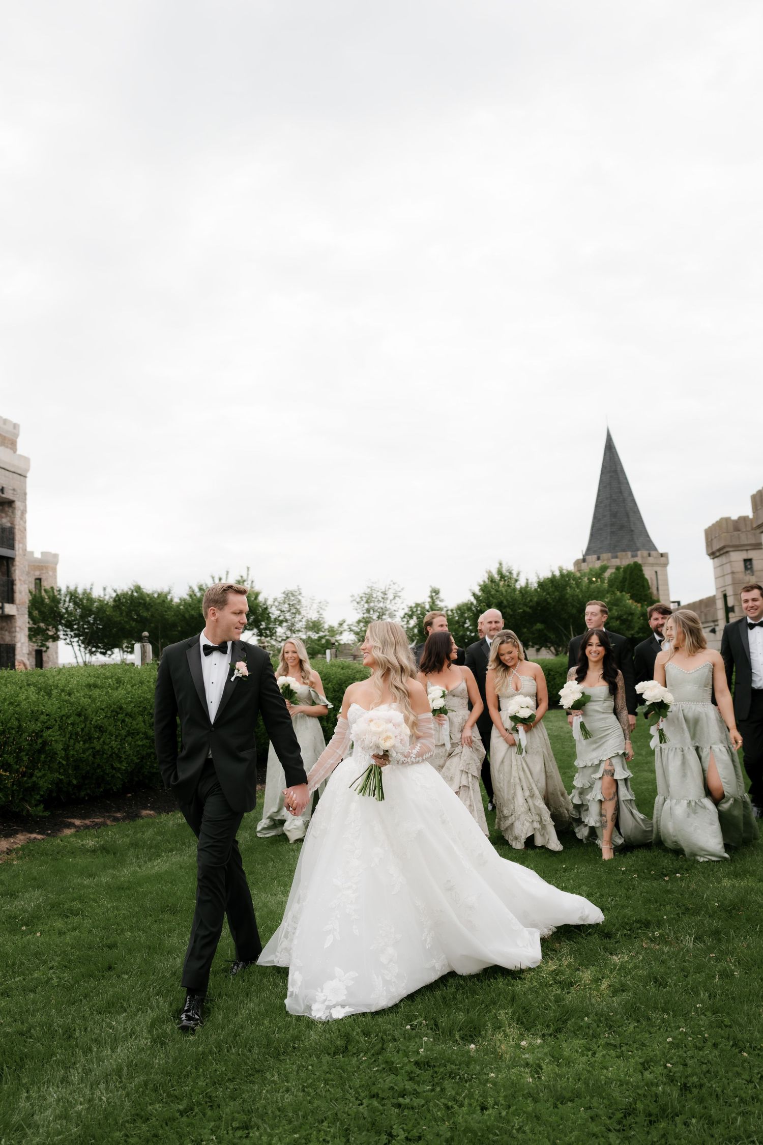 Bride and groom walk hand-in-hand on green lawn with wedding party in elegant attire behind them on a cloudy day.