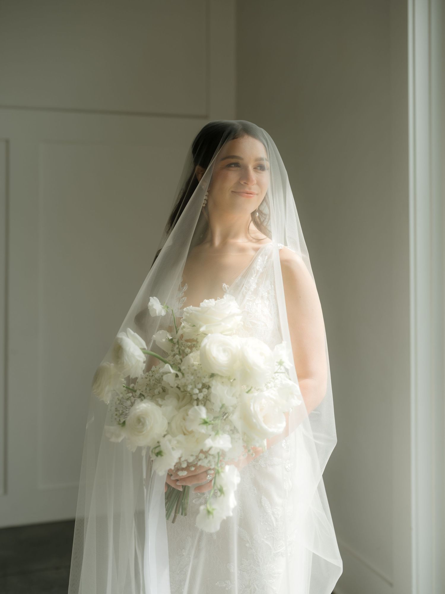 A bride in a white wedding dress and veil holds an elegant white floral bouquet against a light colored wall.
