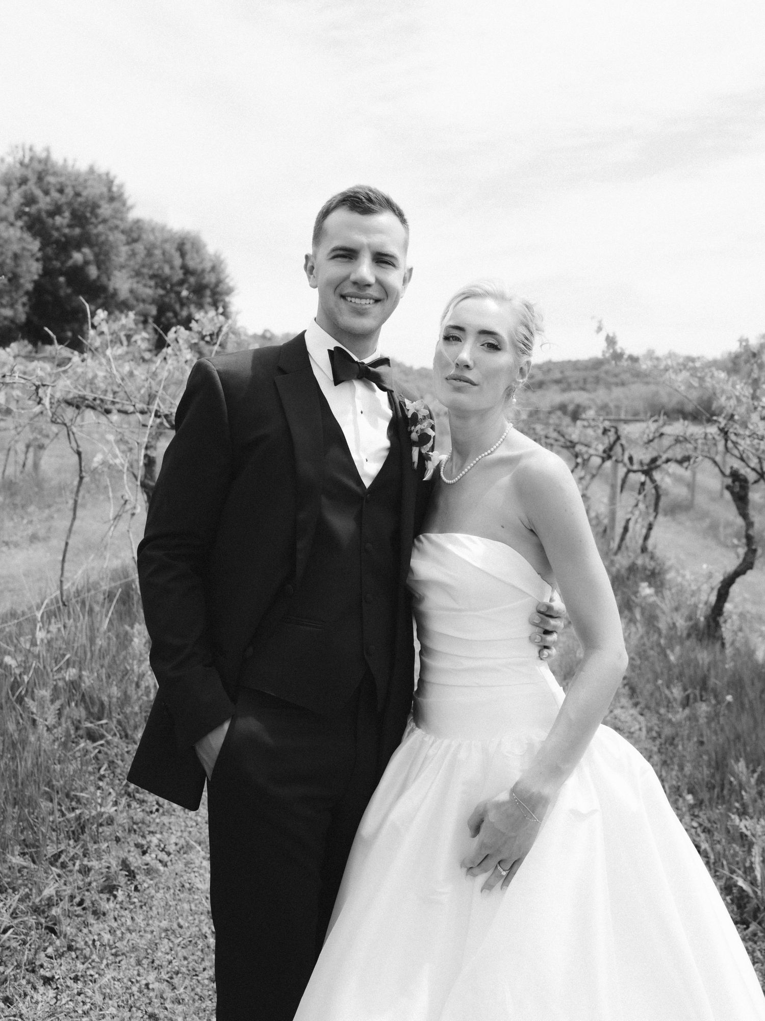 Black and white photo of a smiling groom in a tuxedo and bride in a strapless ball gown posing outdoors at a vineyard.