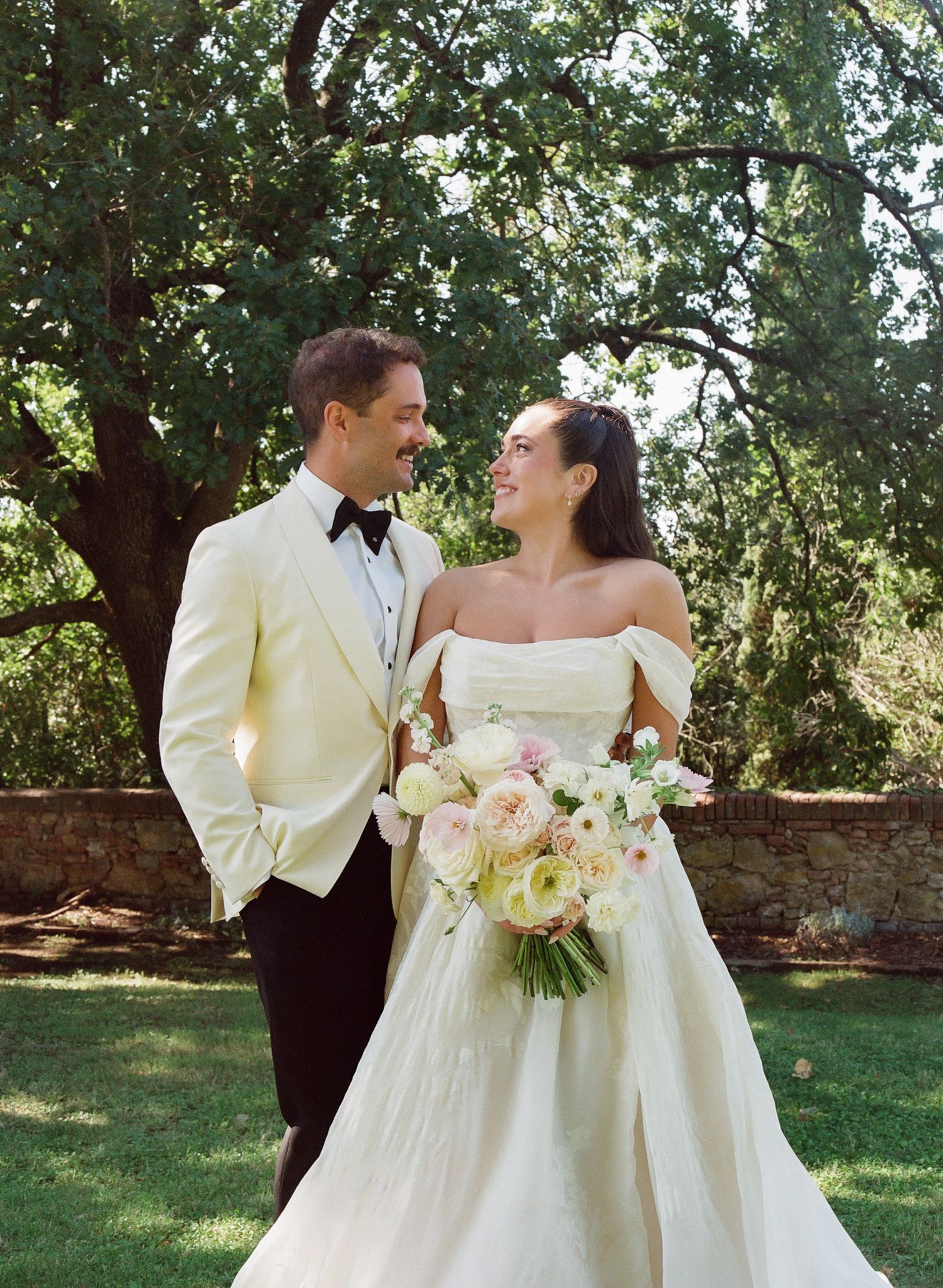 A smiling bride in an off-shoulder gown holds a pink bouquet beside a groom in a cream tuxedo jacket outdoors.