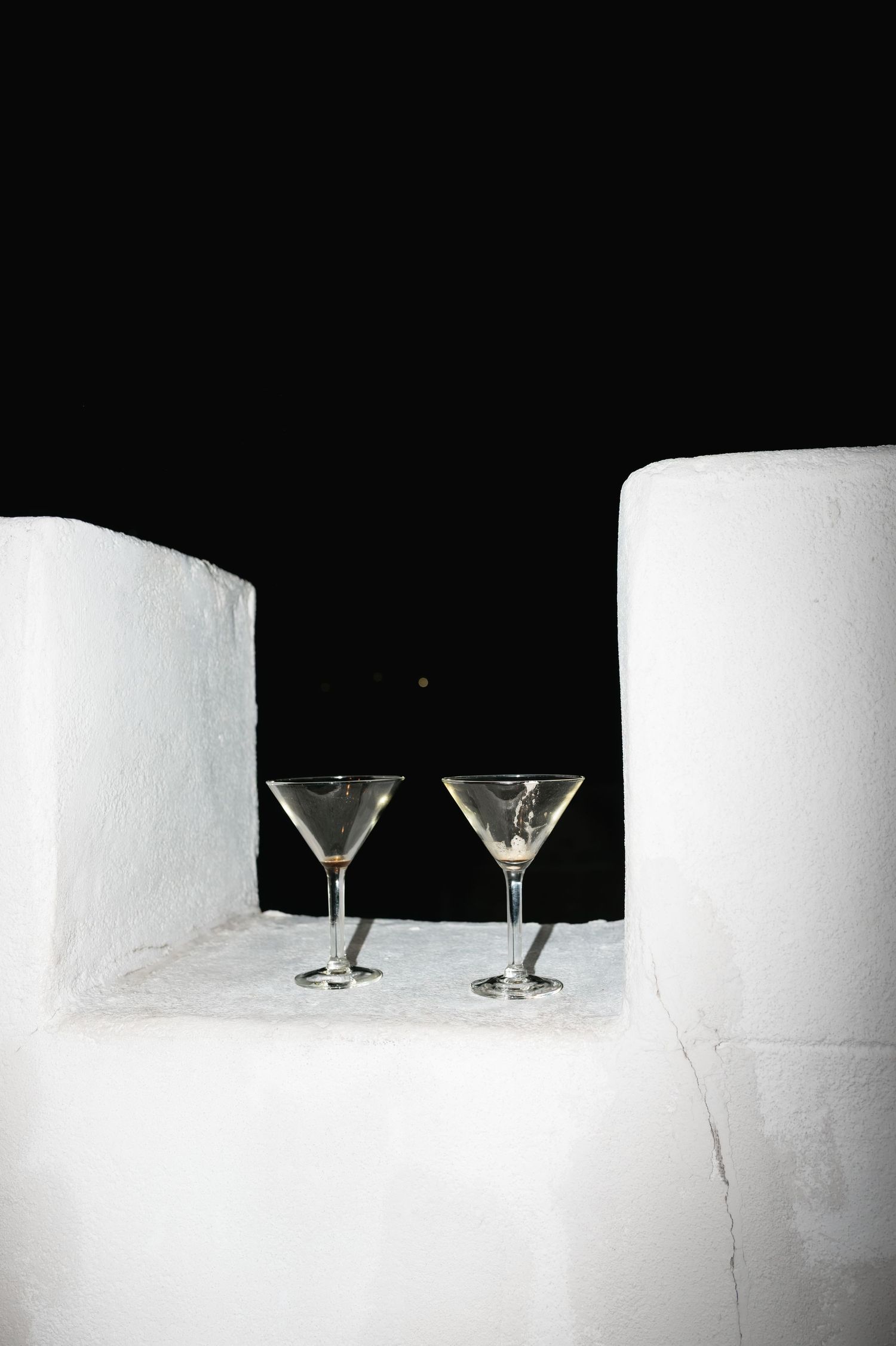 Two martini glasses on a white ledge at night, set against a dark sky in a minimalist outdoor setting.
