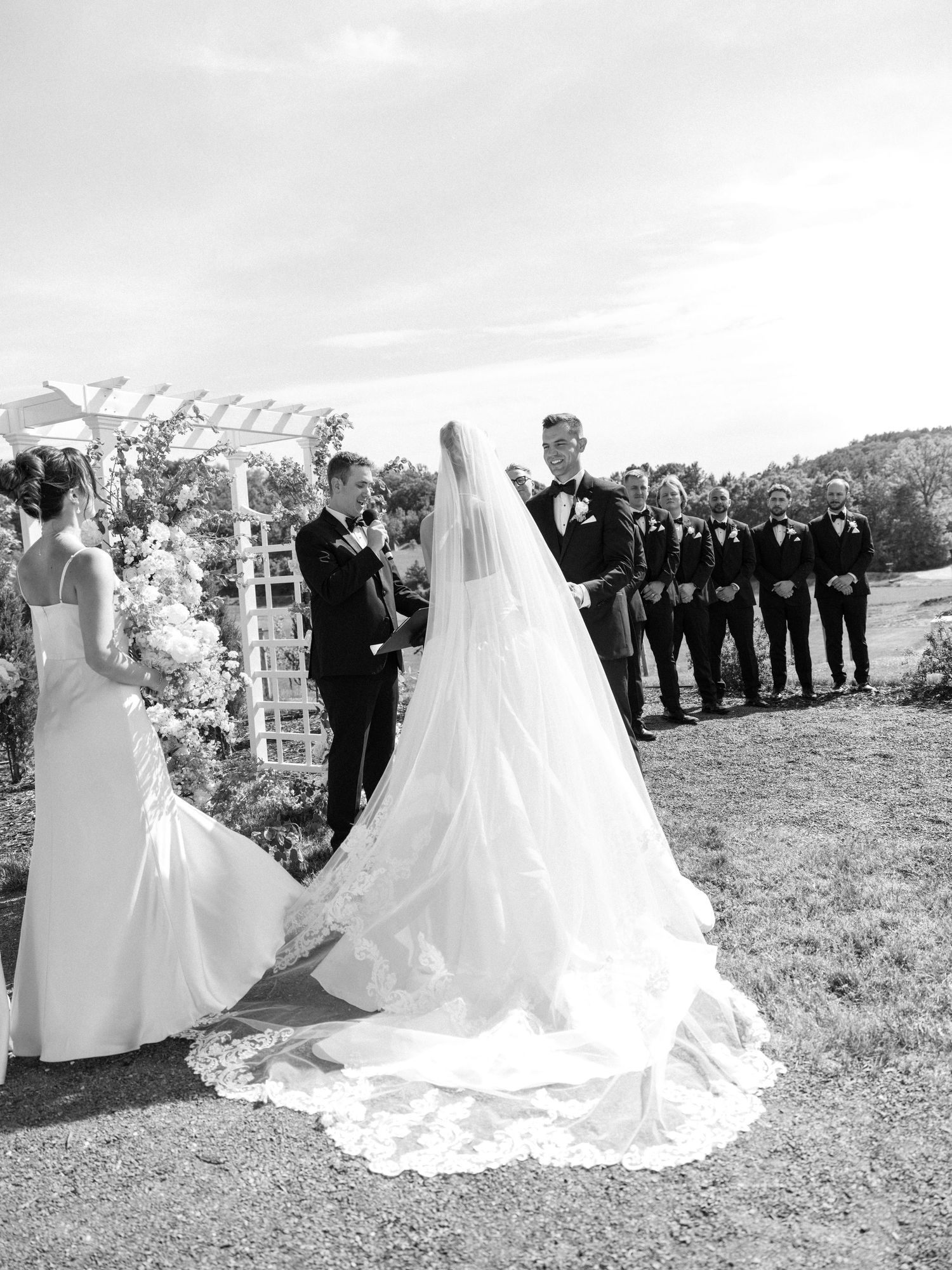 Black and white outdoor wedding ceremony with bride in long veil, groom, officiant, and groomsmen at floral arch.