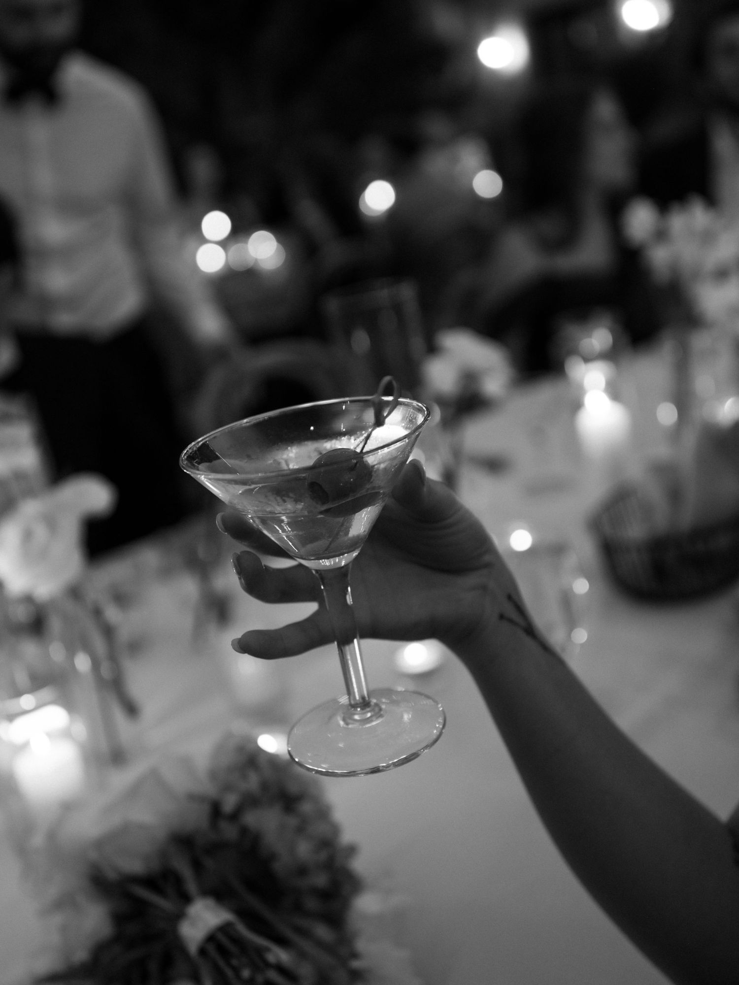 Black and white photo of a hand raising a martini glass at an elegant candlelit wedding reception.
