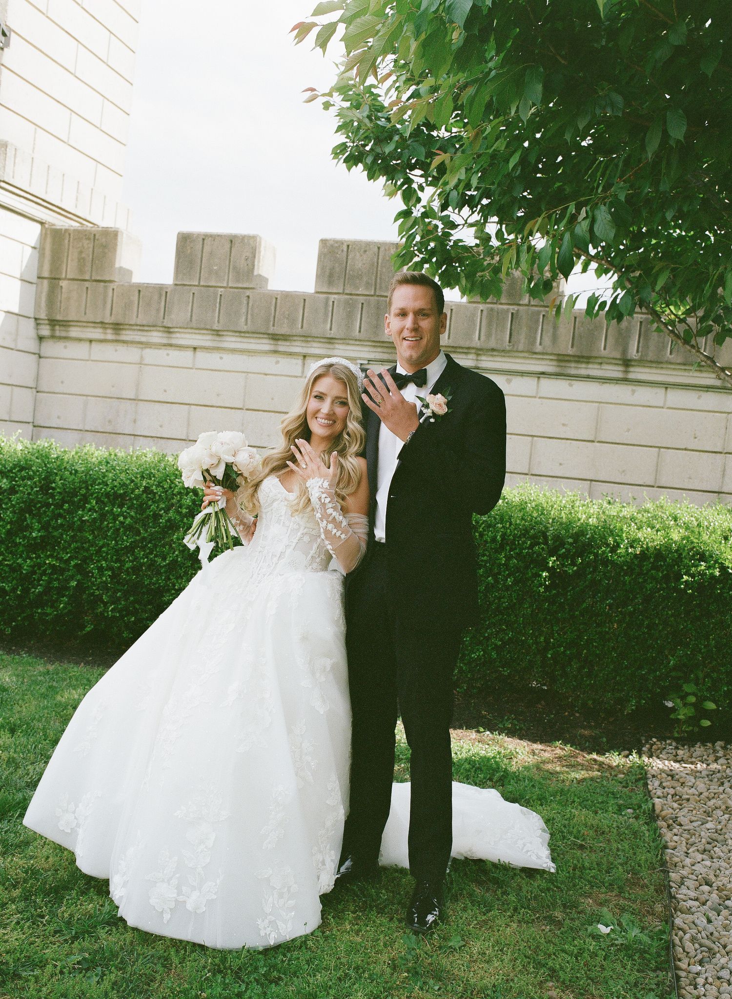 Bride in lace gown holding bouquet stands with groom in black tuxedo showing wedding rings outdoors.