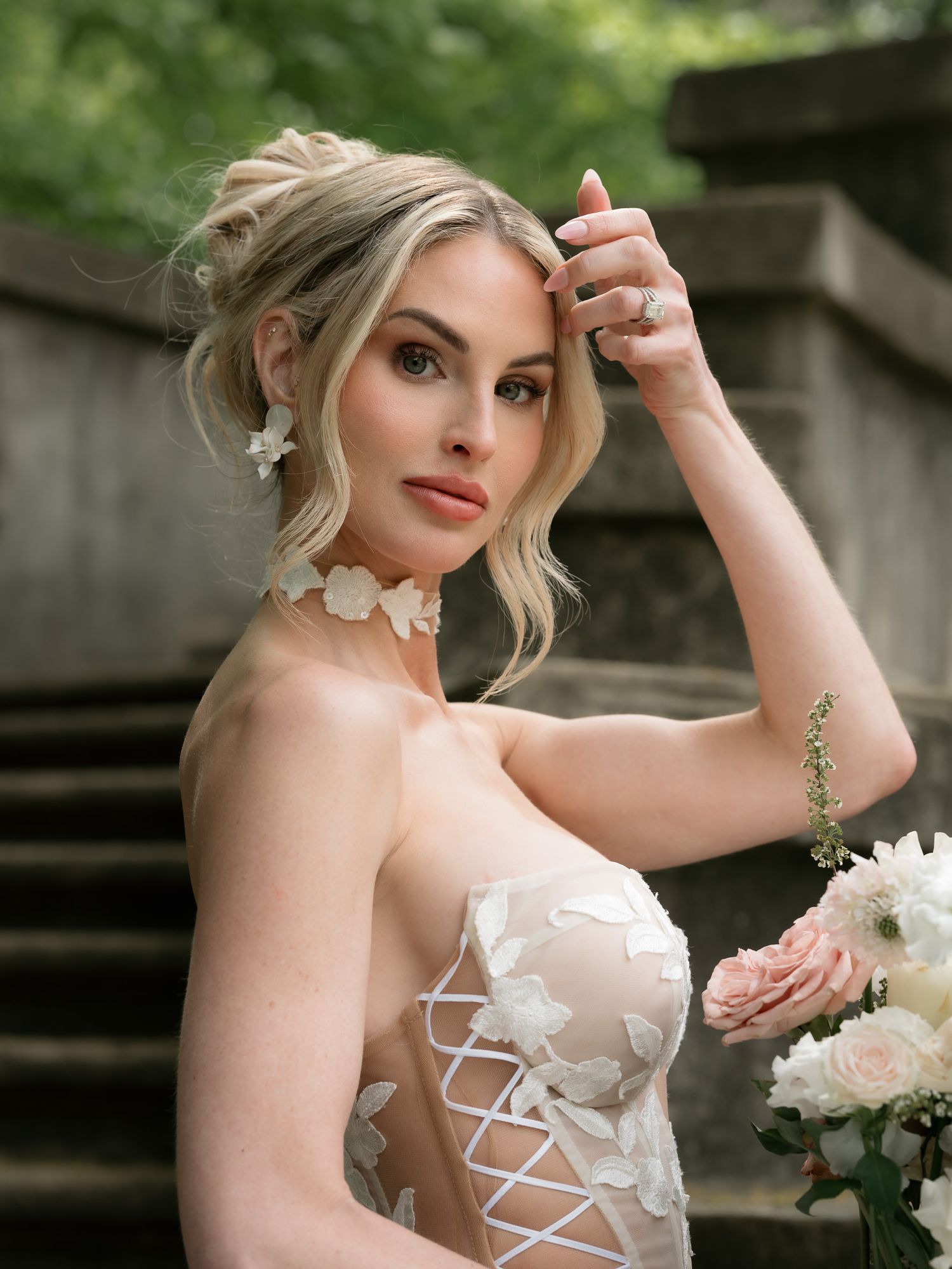 Elegant bride in white floral corset gown with updo hairstyle holding pink bouquet outdoors.