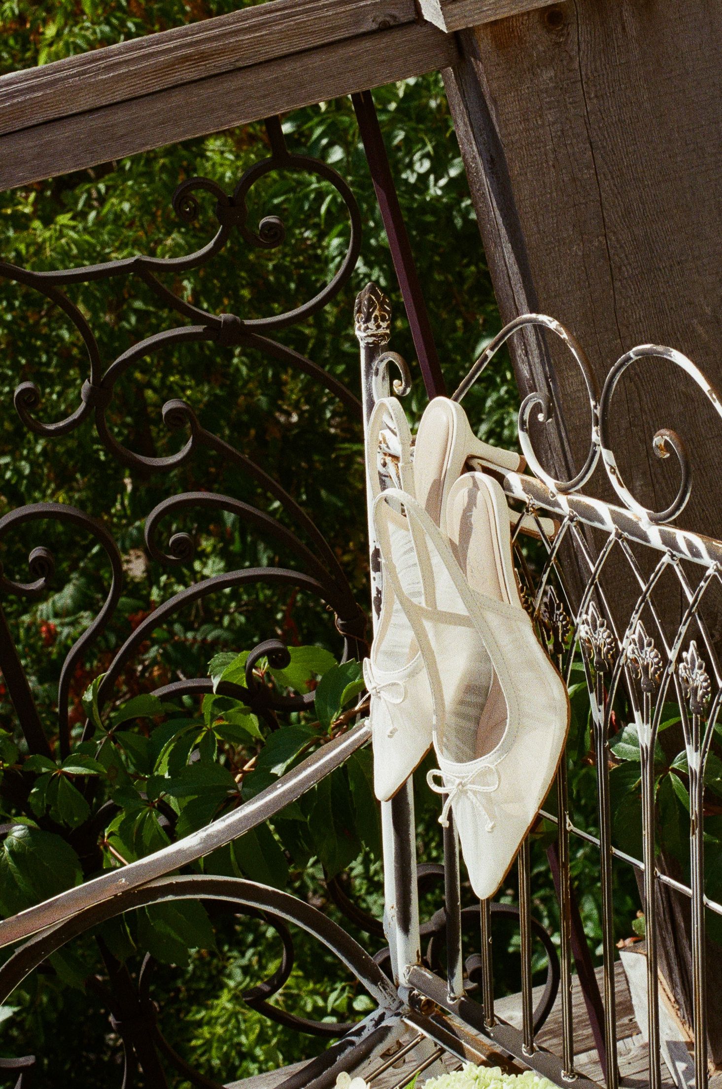 White bridal heels hanging on ornate wrought iron railing with lush green garden background.