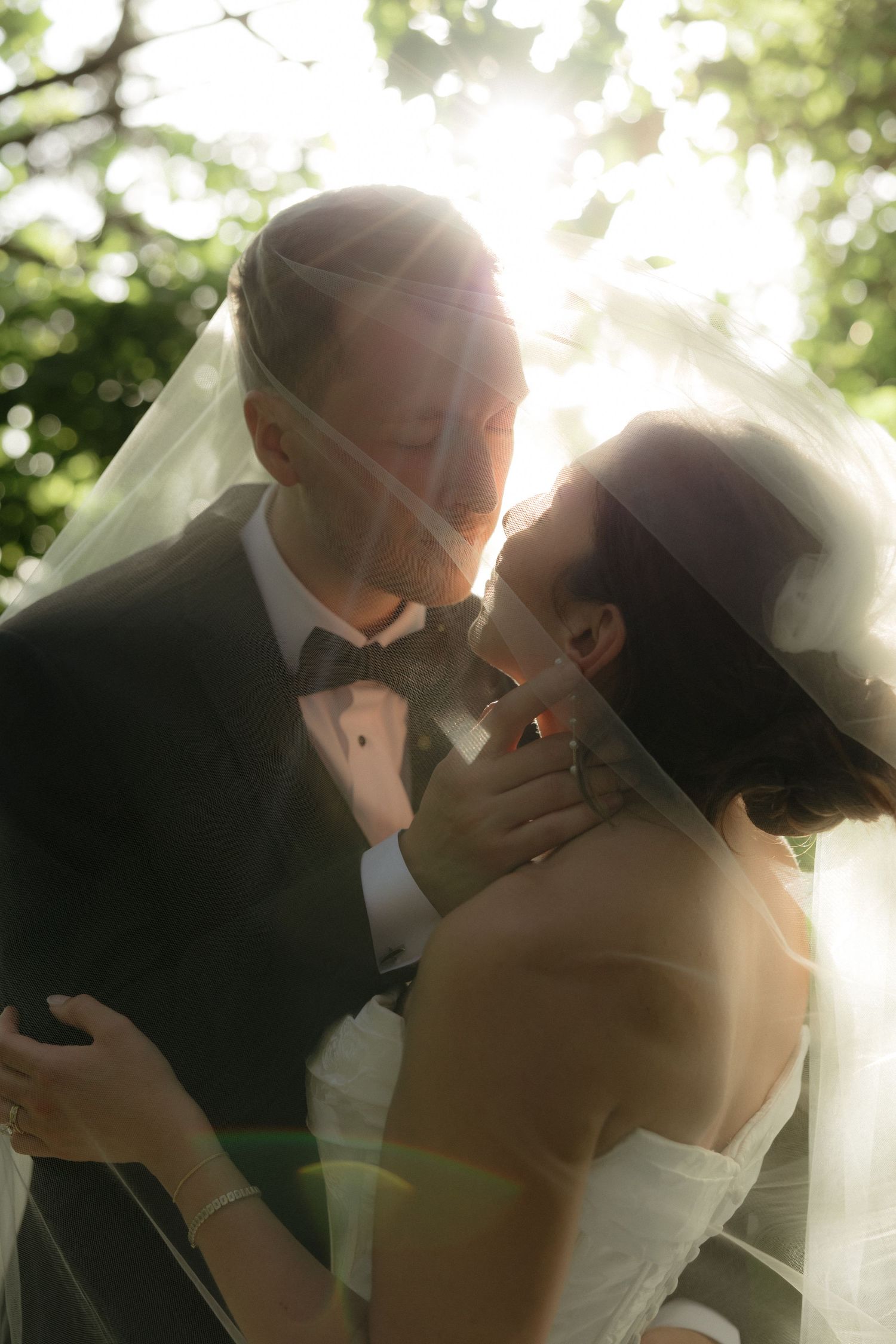 Romantic wedding couple shares an intimate moment under sunlit trees with a flowing bridal veil.