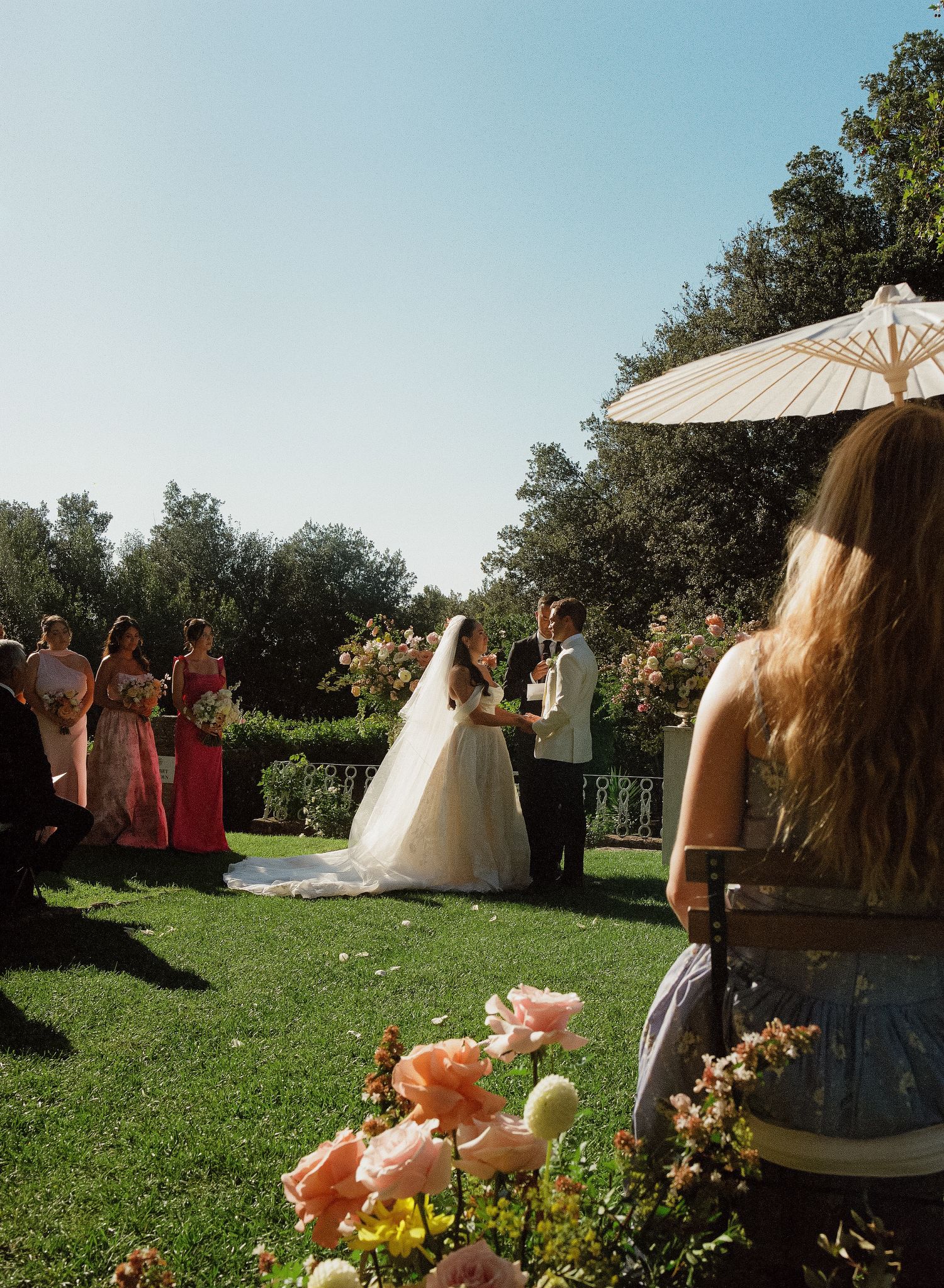 Outdoor garden wedding ceremony with bride and groom at altar, bridesmaids in pink, lush greenery, parasol, and blooming flowers.
