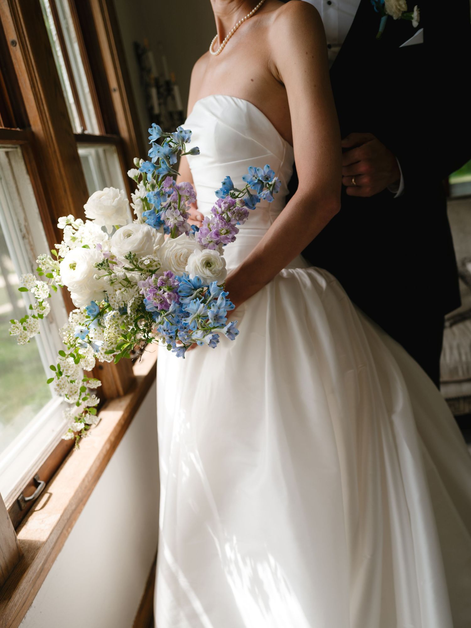 Bride in strapless white ball gown holding blue and white floral bouquet standing by window with groom in black suit.