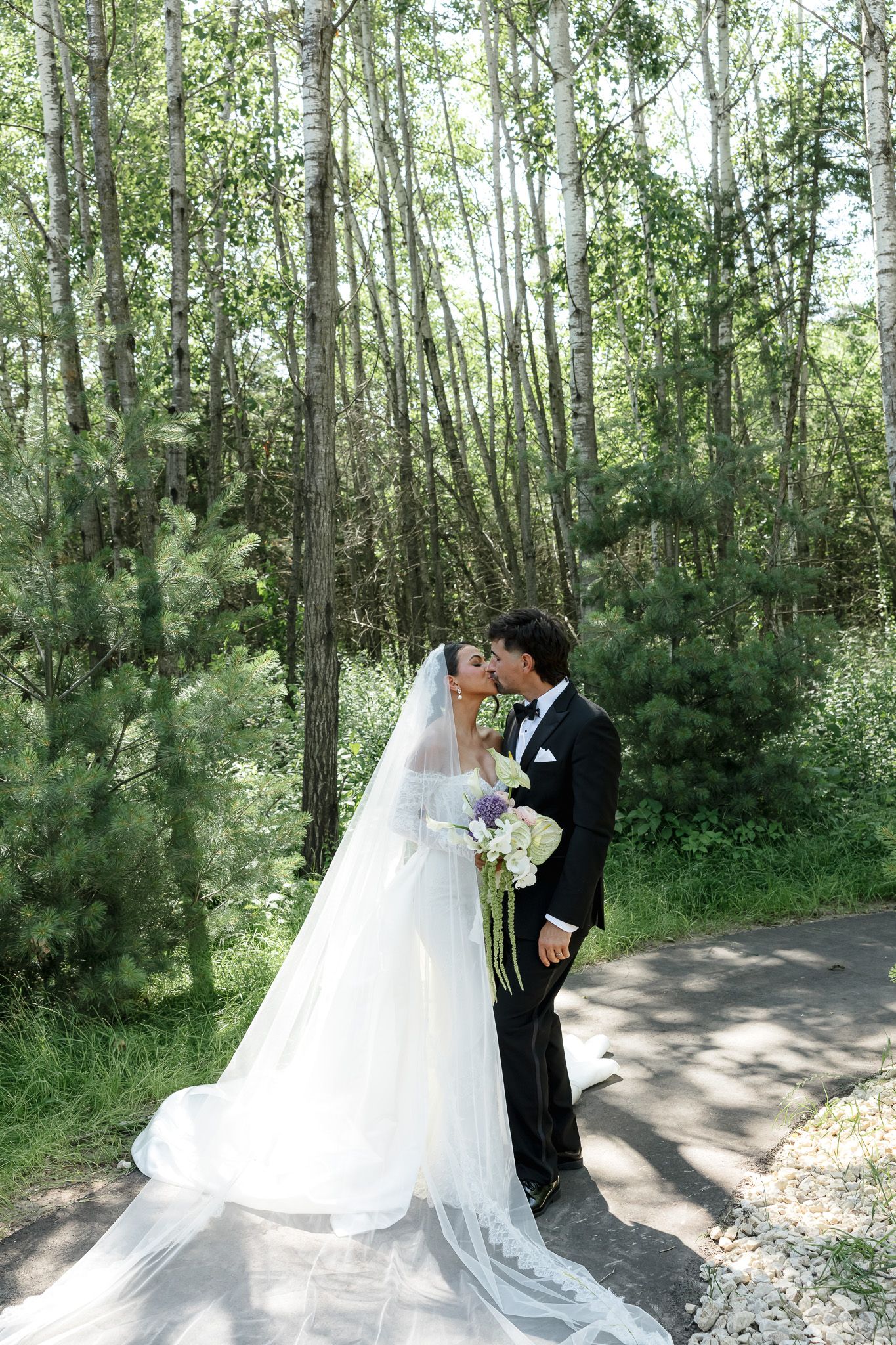 A bride and groom share romantic moments in a sunlit forest path, with her flowing white gown trailing behind them.