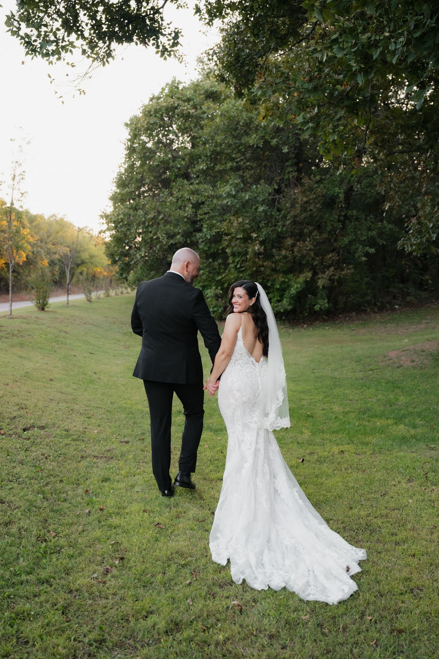 Bride in lace mermaid gown and veil holds hands with groom in black tuxedo while walking on green lawn near trees.