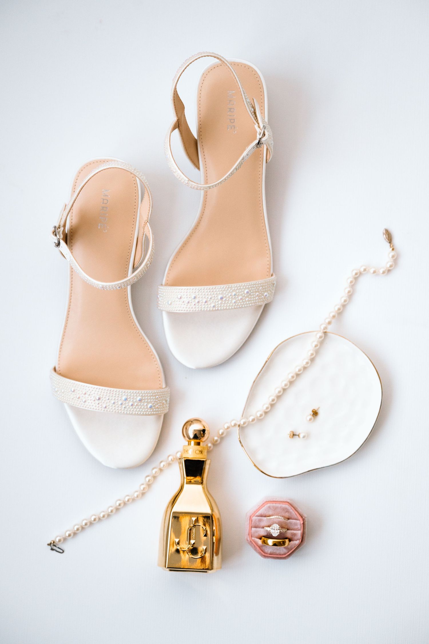 White bridal sandals with pearl necklace, gold perfume bottle, pink ring box, and earrings on white background.