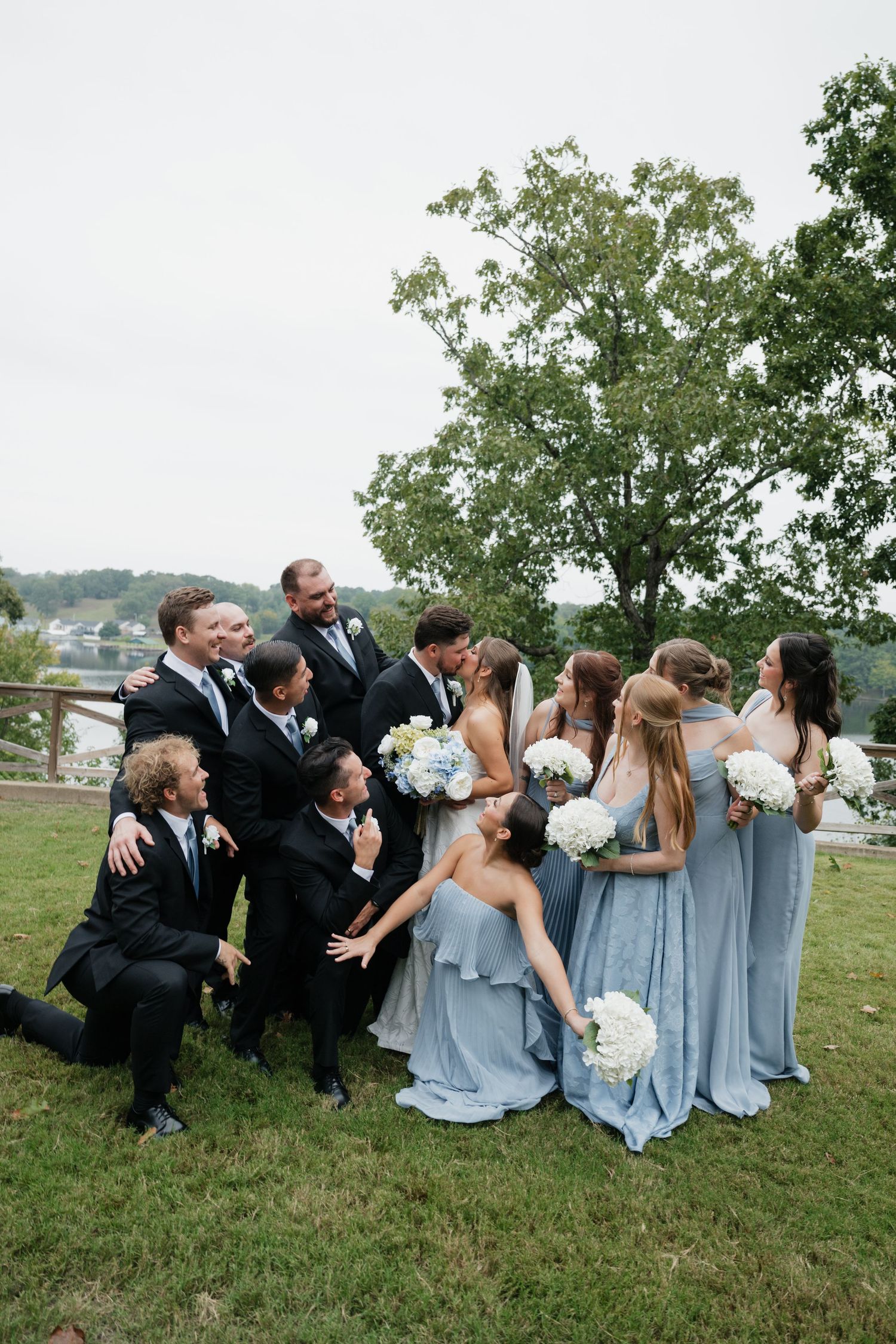 Wedding party poses outdoors, bridesmaids in blue gowns holding white bouquets surround kissing couple on green lawn.