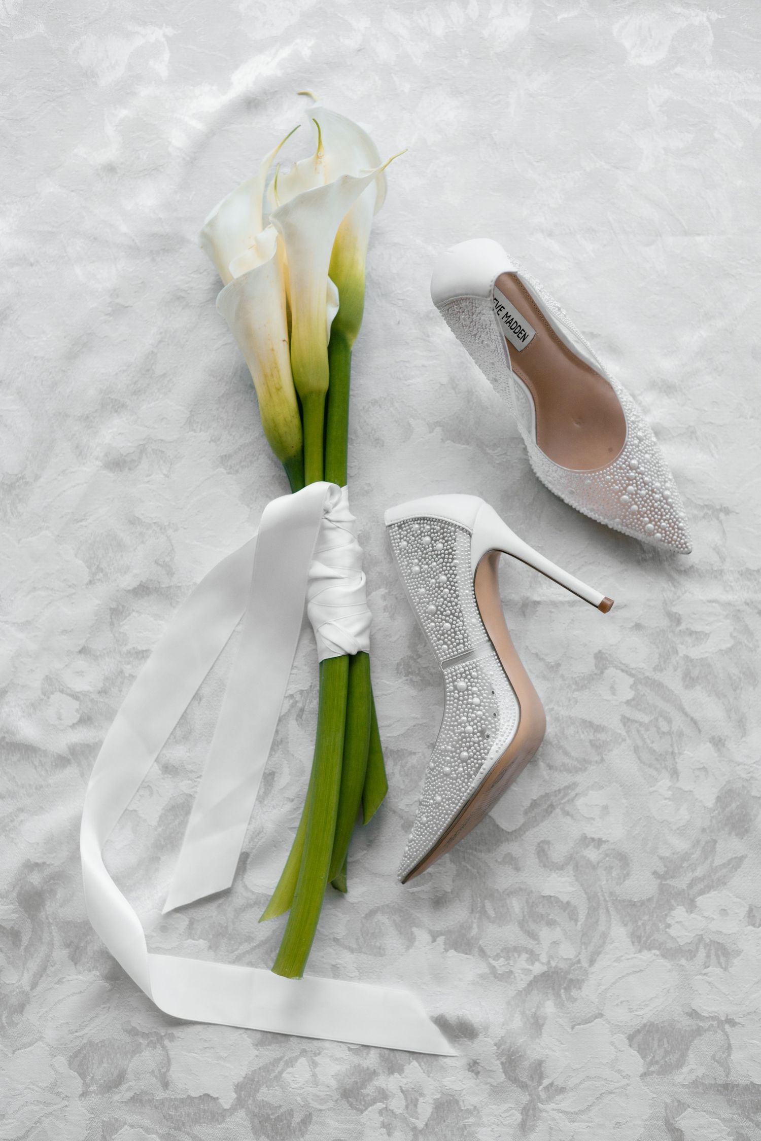White bridal heels with crystal embellishments beside a calla lily bouquet tied with white ribbon on marble.