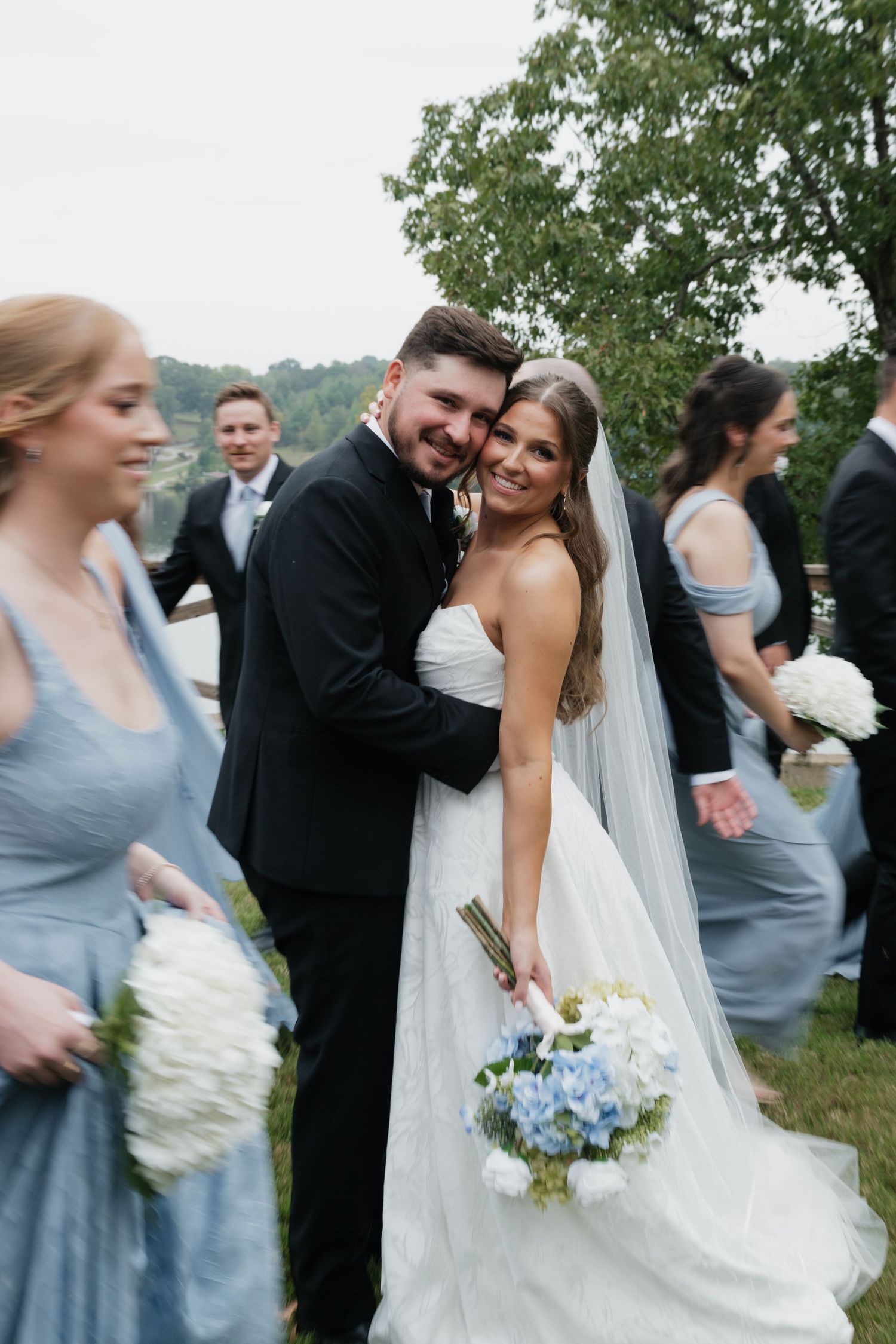 Bride and groom smile together at outdoor wedding, surrounded by bridesmaids in blue dresses holding white bouquets.