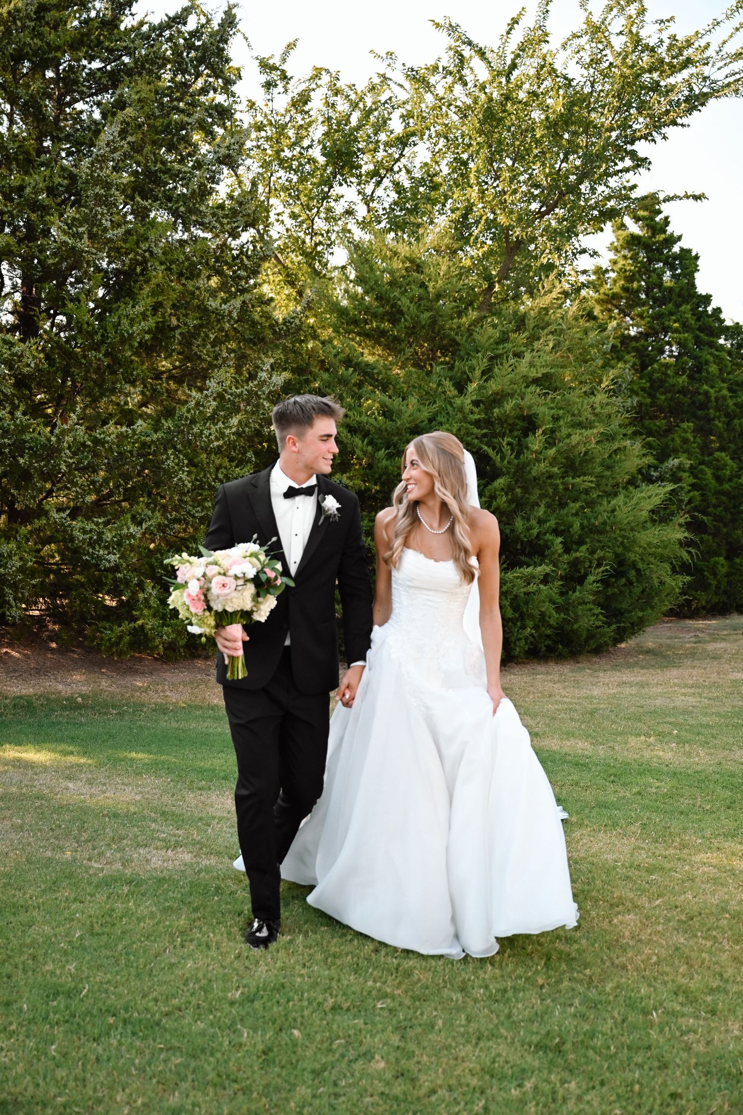 A bride and groom walk hand-in-hand on green grass, smiling at each other, with lush trees in the background.