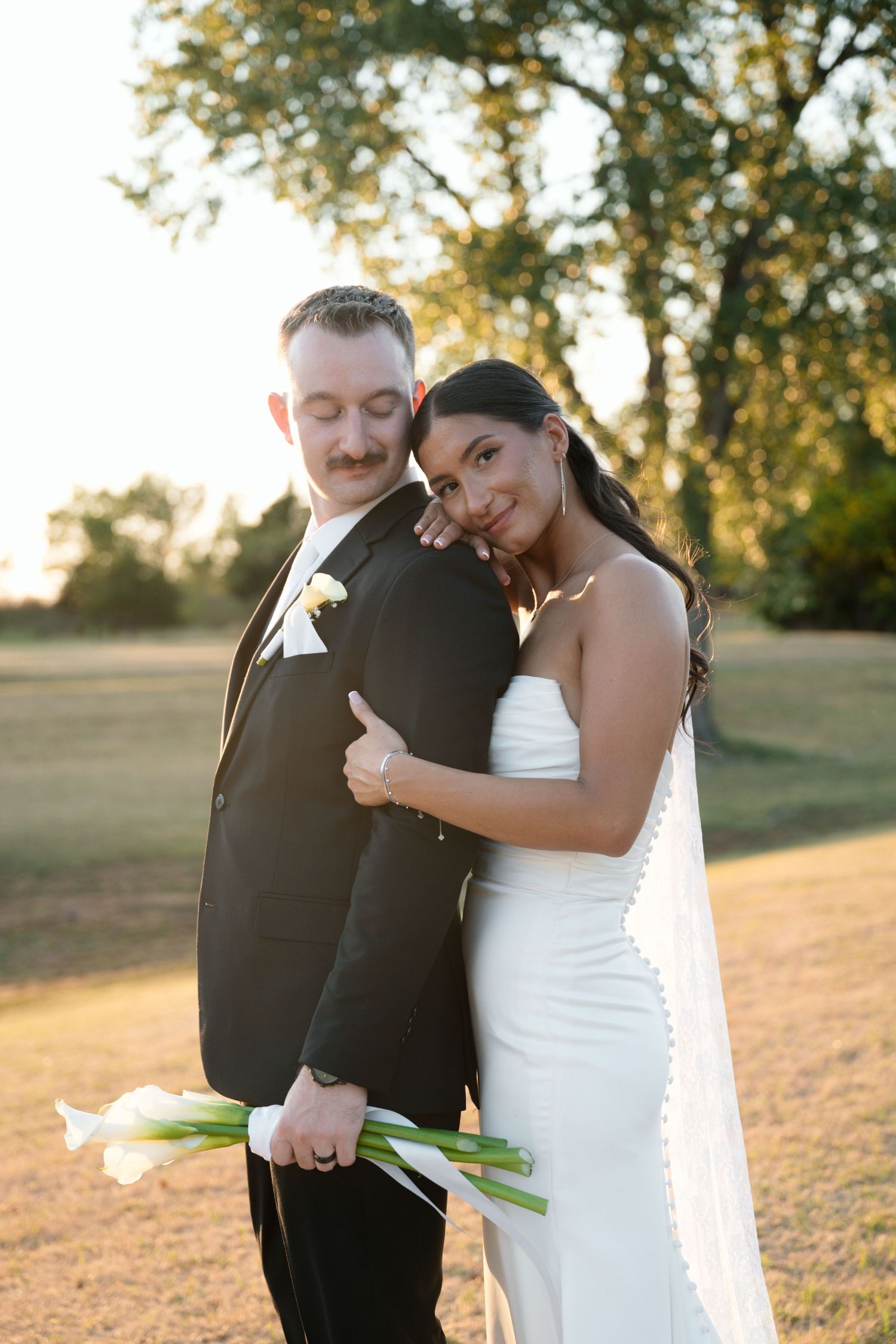 Bride in white gown hugs groom in black suit outdoors at golden hour, holding white calla lilies.