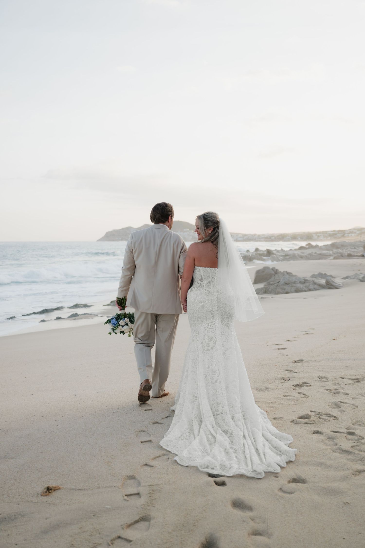 Bride in a lace wedding gown and groom in a tan suit walk hand in hand along a sandy beach at sunset, leaving footprints near the ocean.