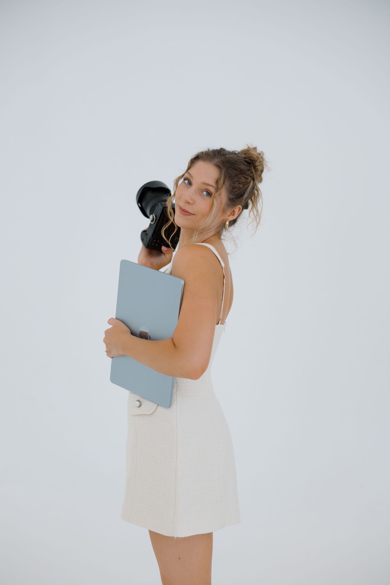 Smiling woman in a white sundress with hair in a messy bun holds a camera and a blue-gray folder against a clean white studio background.