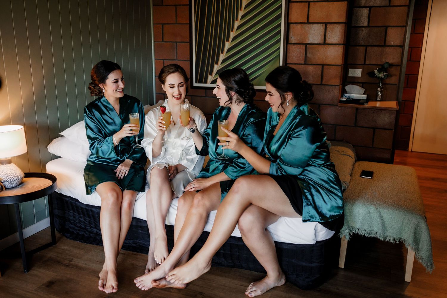 Four bridesmaids in teal silk robes enjoying drinks together during wedding preparations.