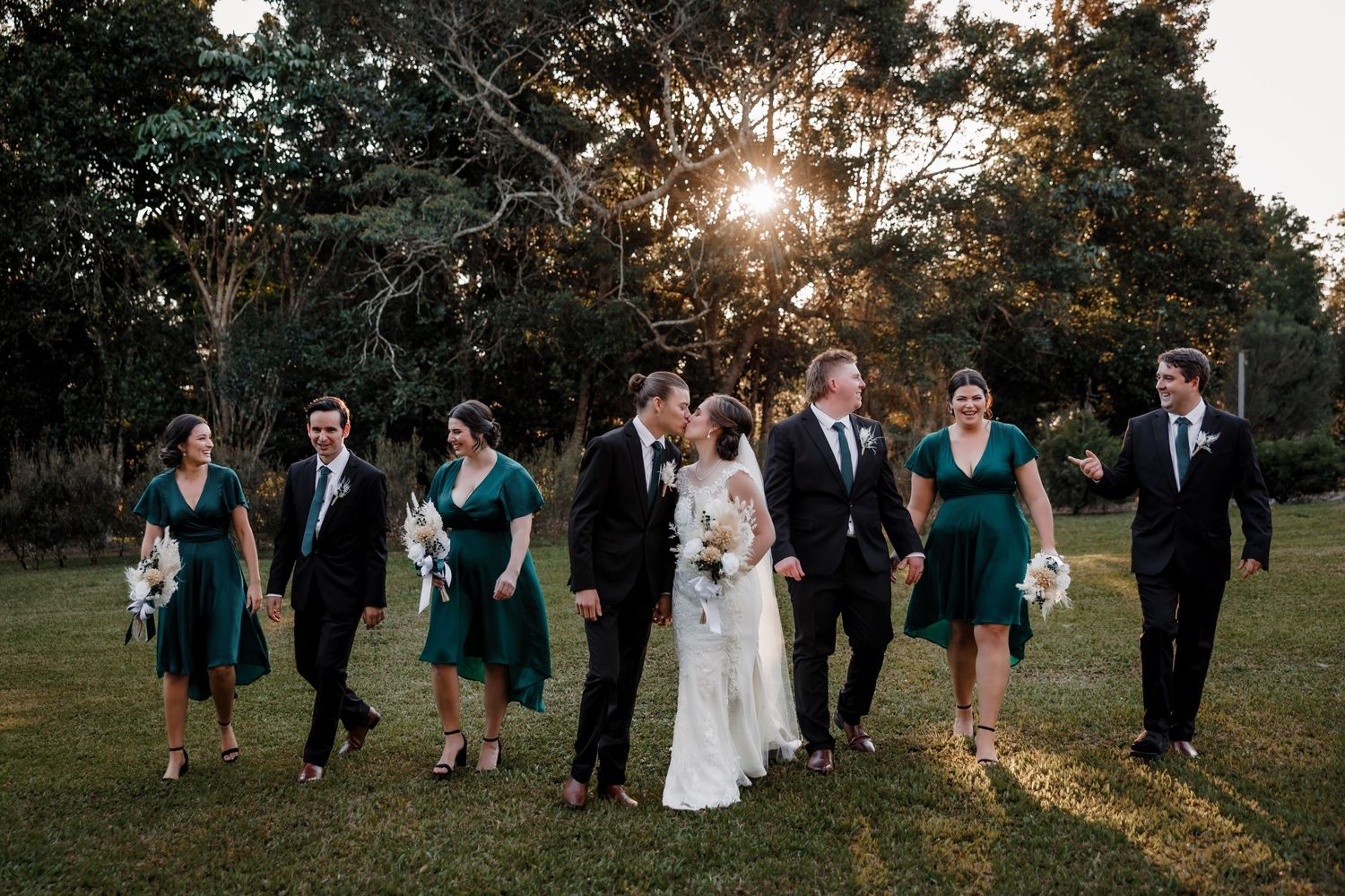 Wedding party in emerald dresses walking through a sunlit garden at golden hour.