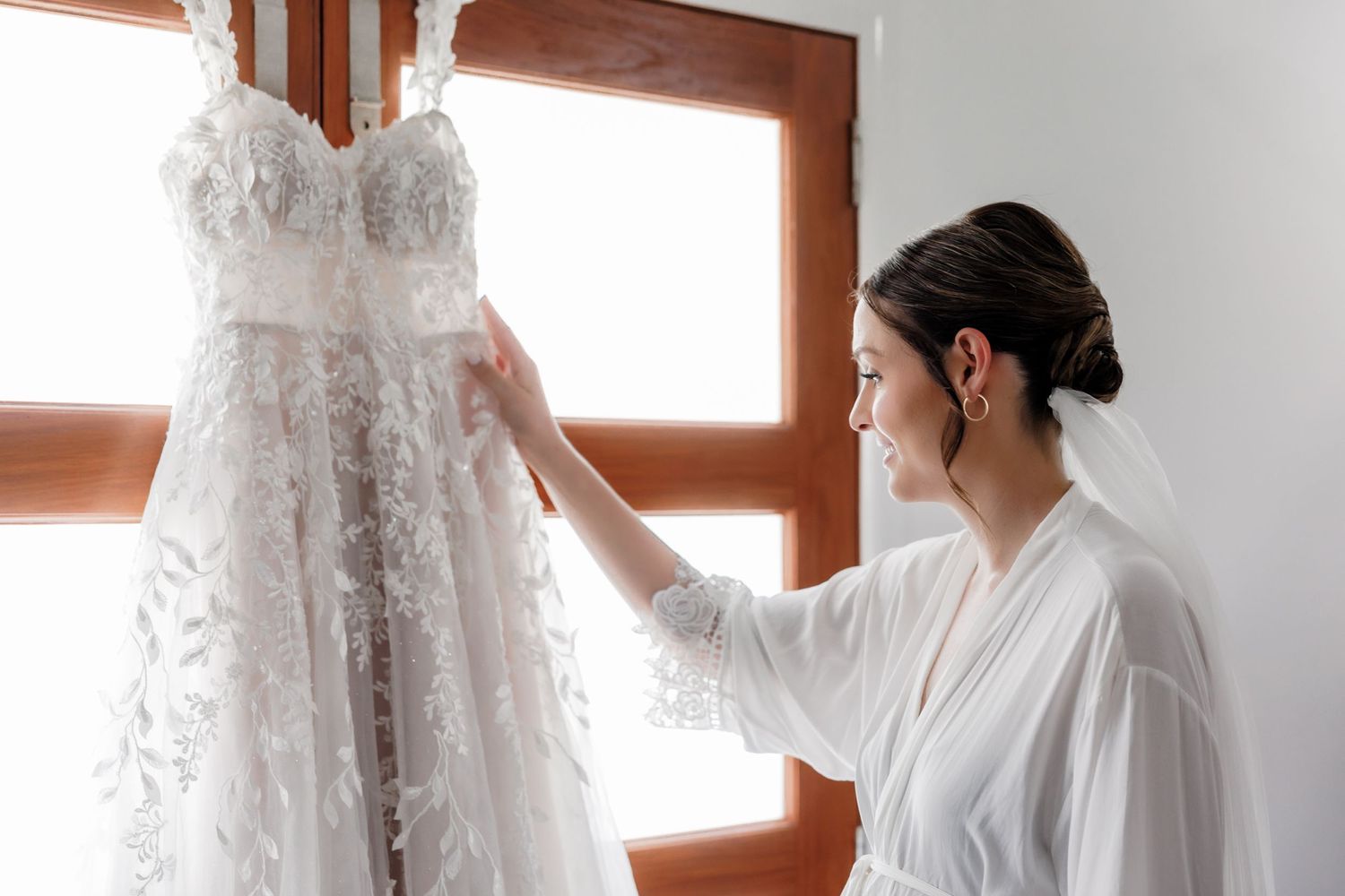 Elegant lace wedding dress hangs in window light during bridal preparation.