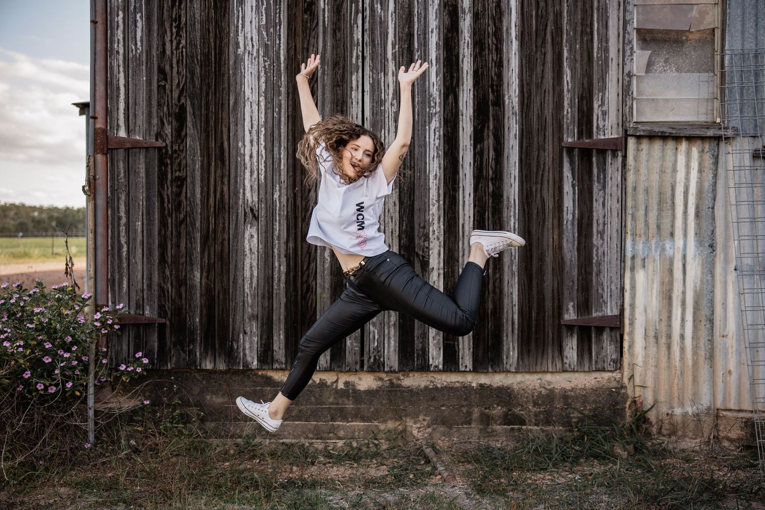 A joyful person in a white shirt and jeans jumps playfully in front of a rustic wooden barn wall.